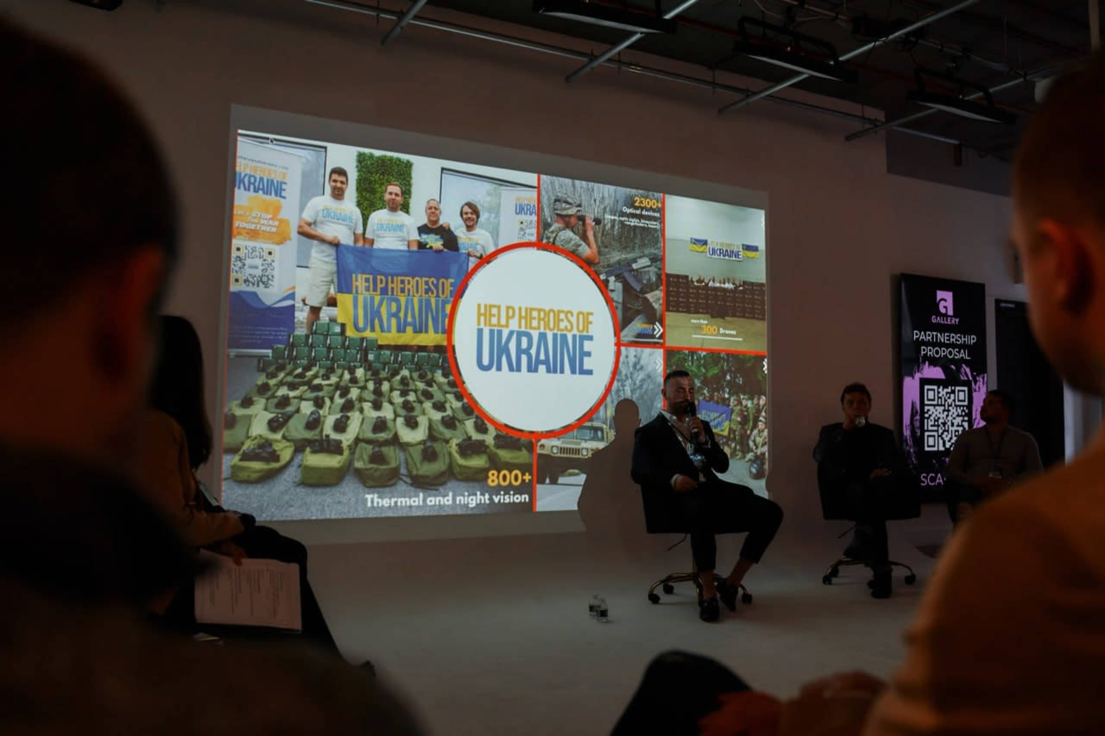 A presentation is taking place in a modern venue, featuring a large screen displaying the "Help Heroes of Ukraine" campaign alongside images of various support efforts.