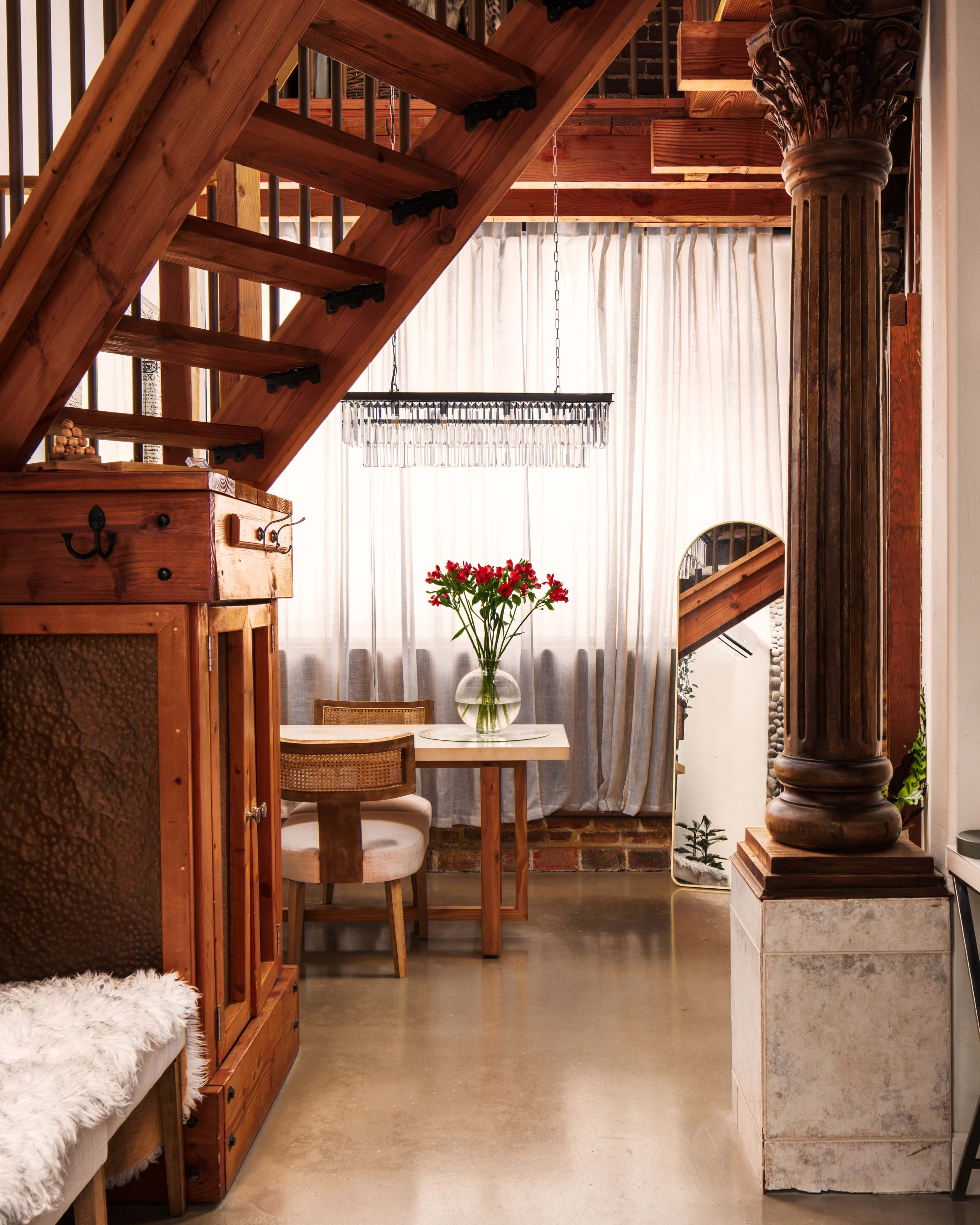 The image shows a stairway made of wood leading to a second level, with a dining area featuring a round table and a vase of flowers beside a large window draped with sheer curtains.