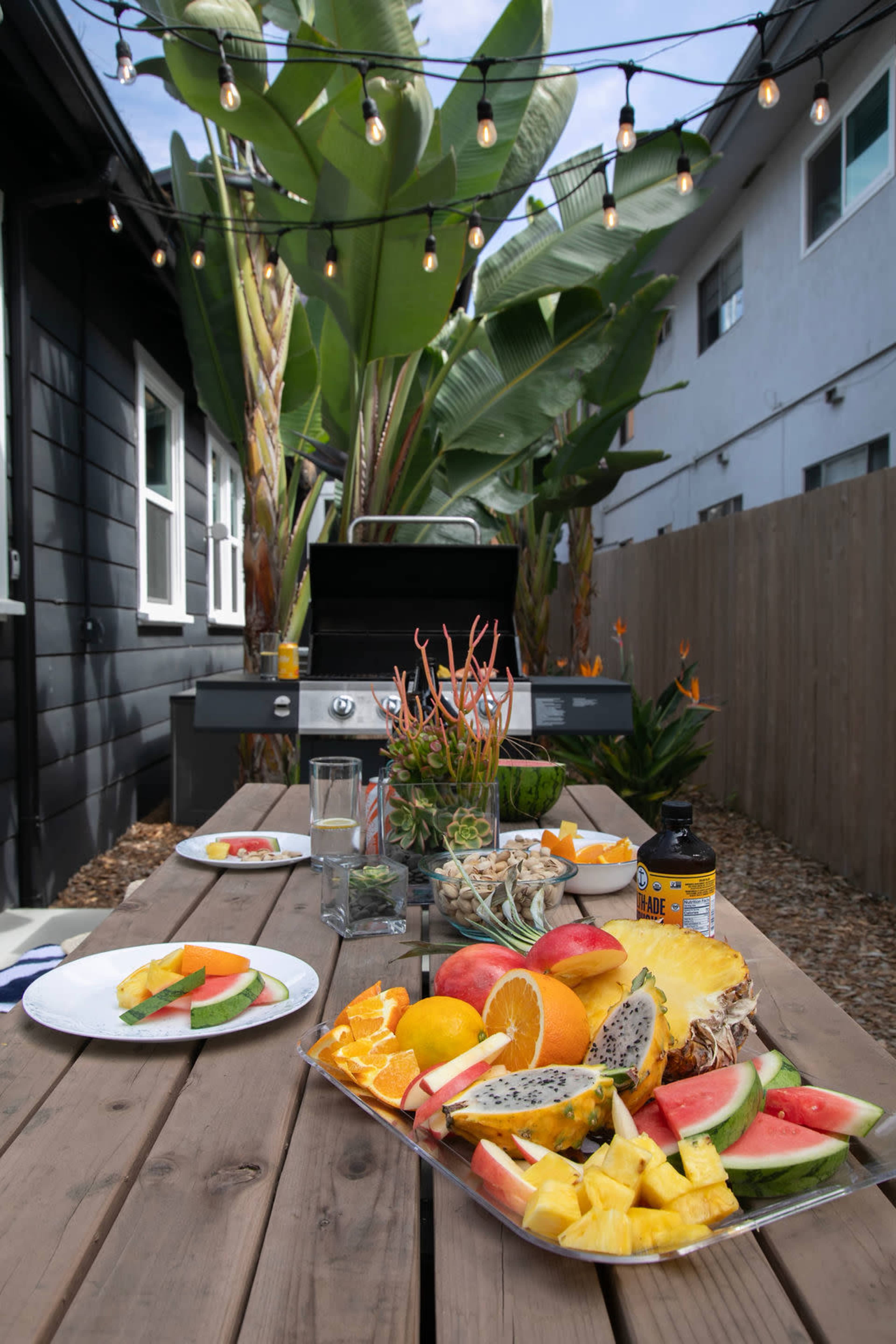 A wooden table is set with an array of fresh fruits and a grill is visible in the background, surrounded by palm trees and string lights.
