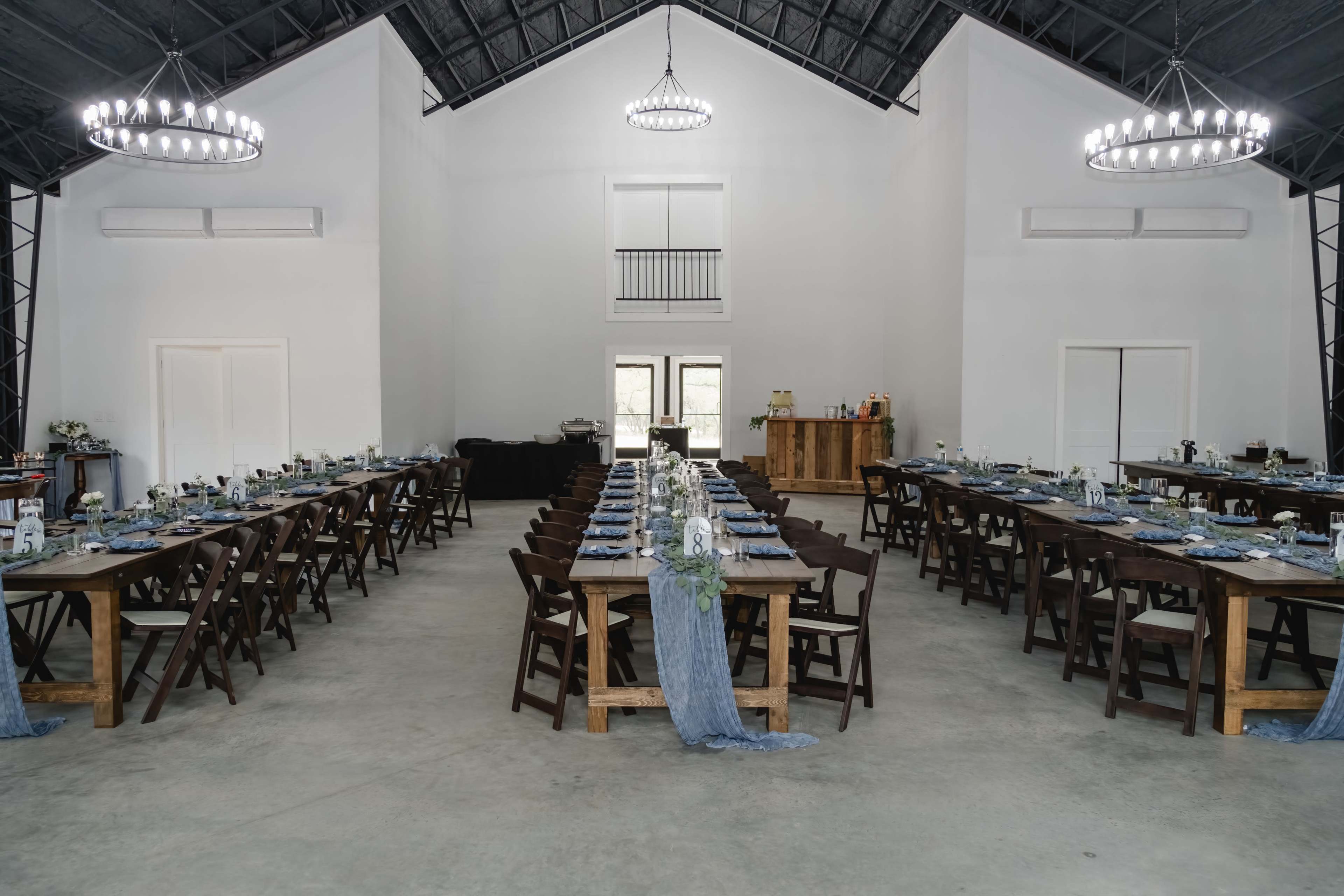 The image shows a spacious interior with two long dining tables set for an event, surrounded by white walls and a high ceiling with chandeliers.