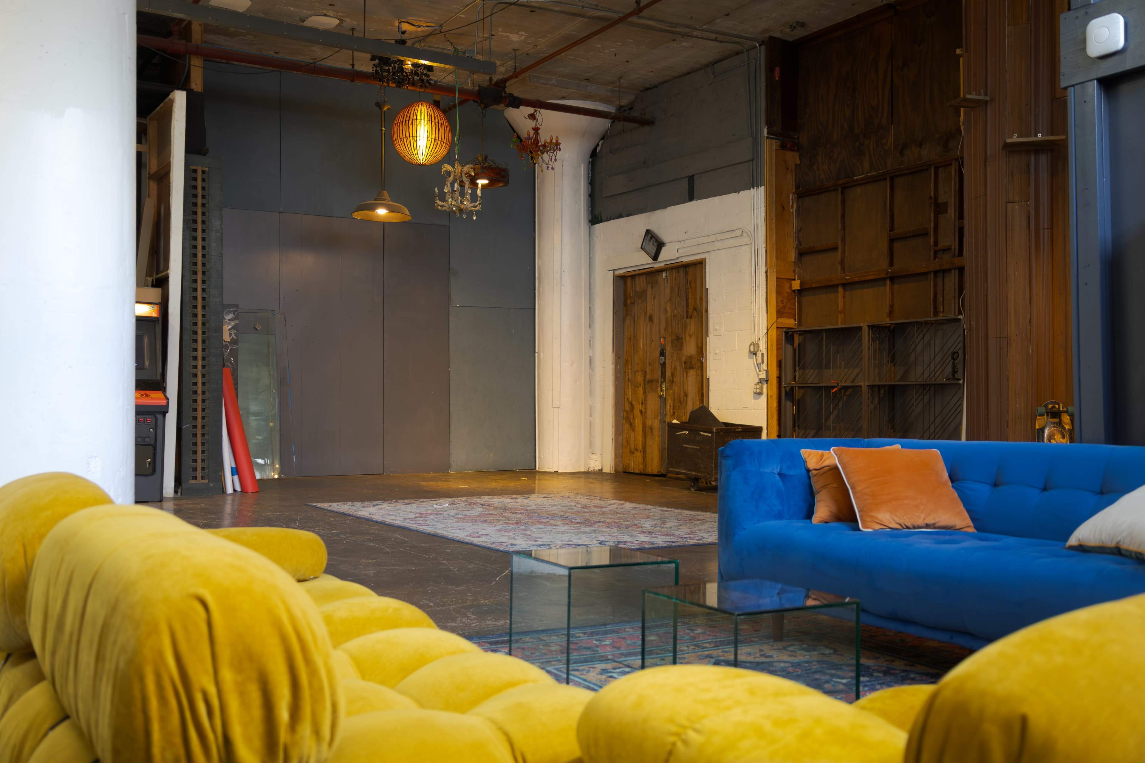The image shows a spacious interior with a blue couch and a yellow tufted sofa facing a glass coffee table in a loft-like setting with various textures and lighting fixtures.