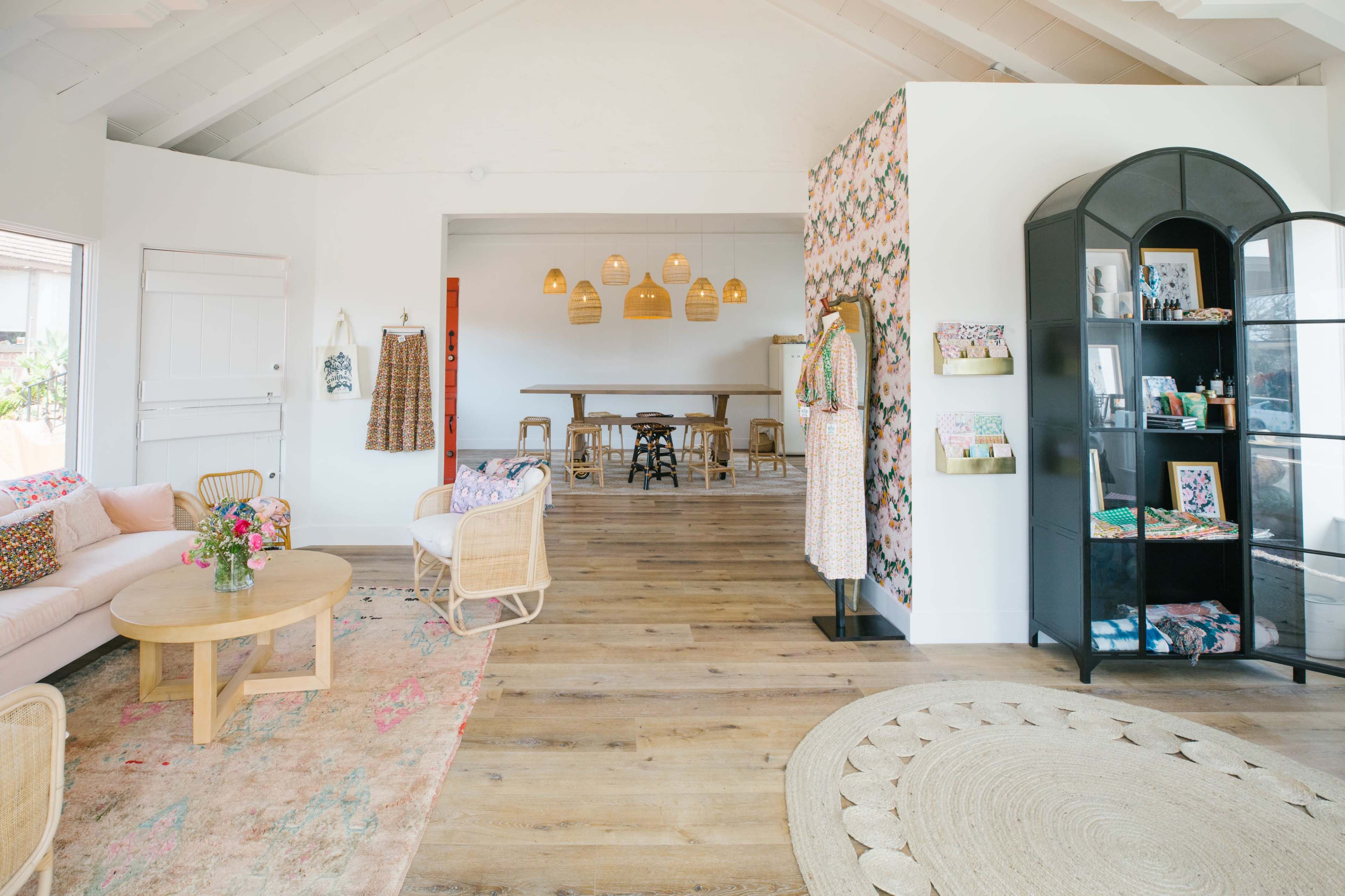 A bright, open interior space featuring a seating area with a sofa and a coffee table, a dining area with a long table, and a display shelf against a floral wall.