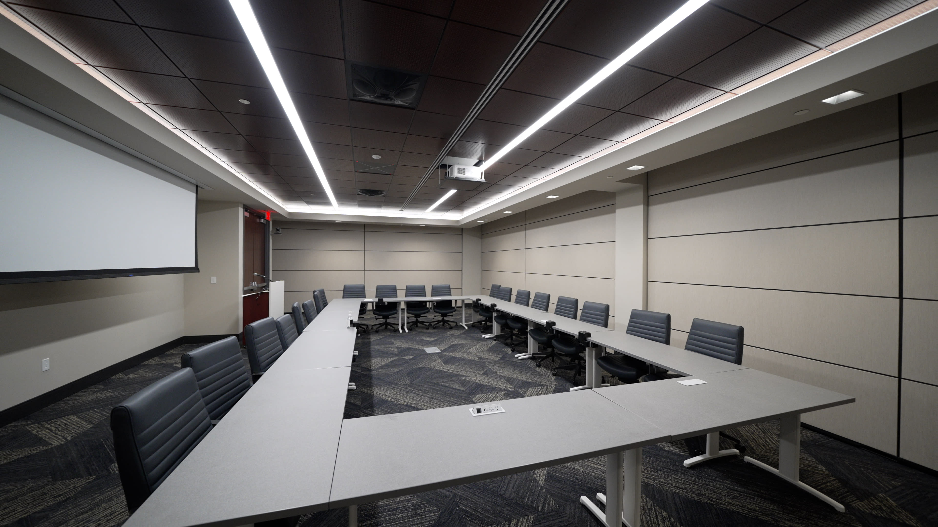 A modern conference room arranged with a long table surrounded by black office chairs and equipped with a projector screen.