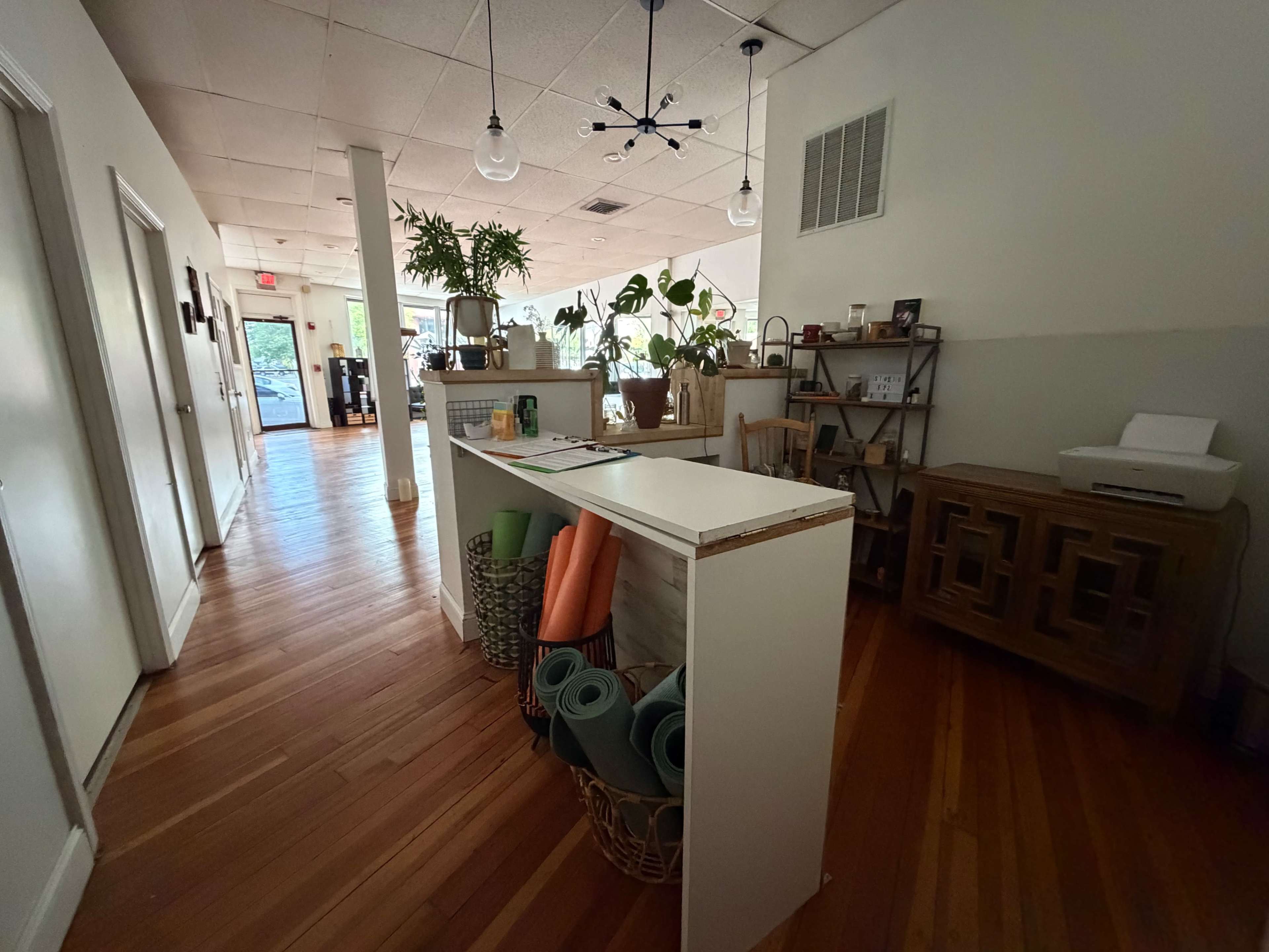 The image shows a spacious interior of a welcoming reception area with wooden floors, plants, and yoga mats on display.