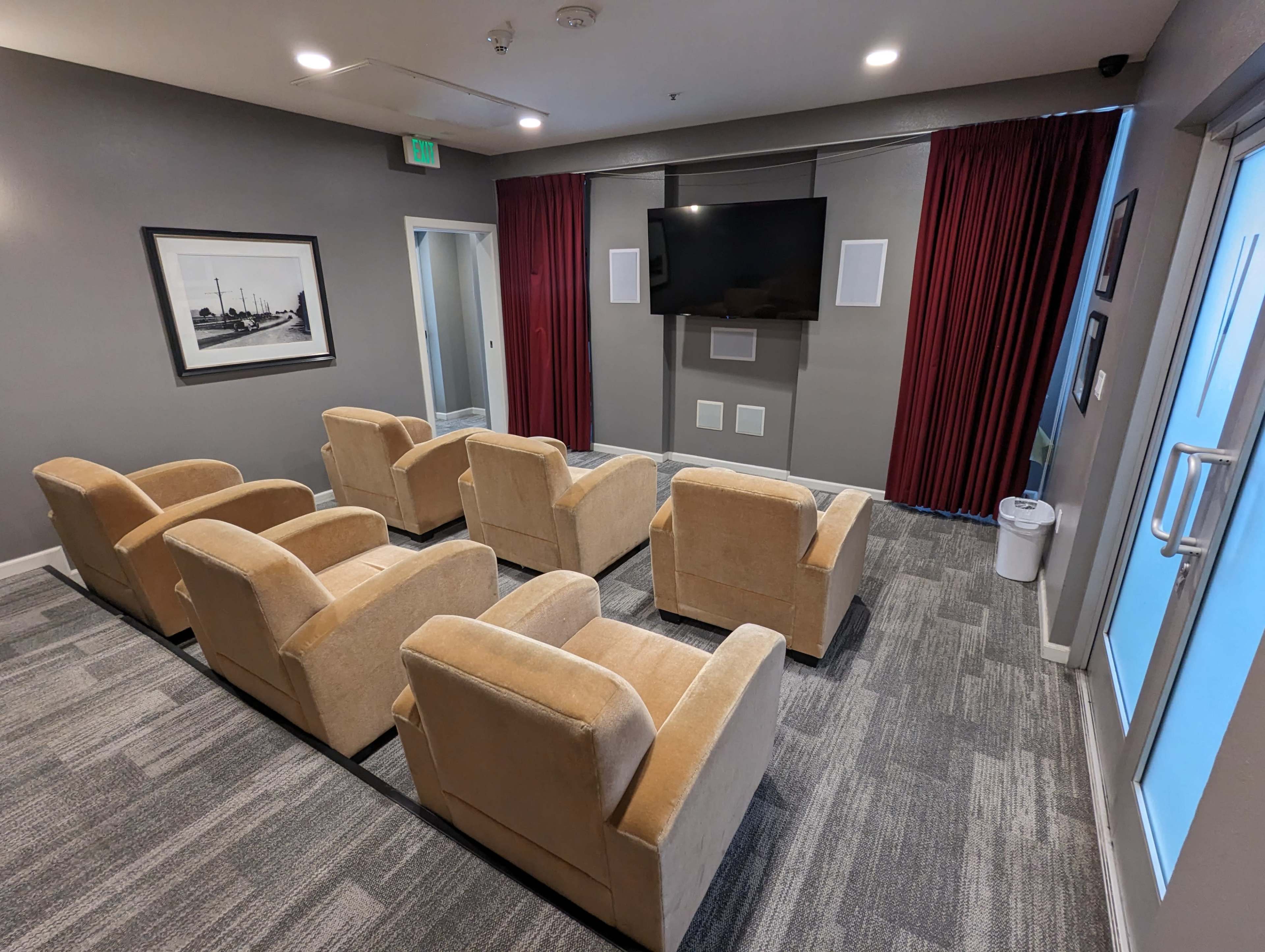 A small screening room with six beige chairs facing a television mounted on the wall, flanked by red curtains.