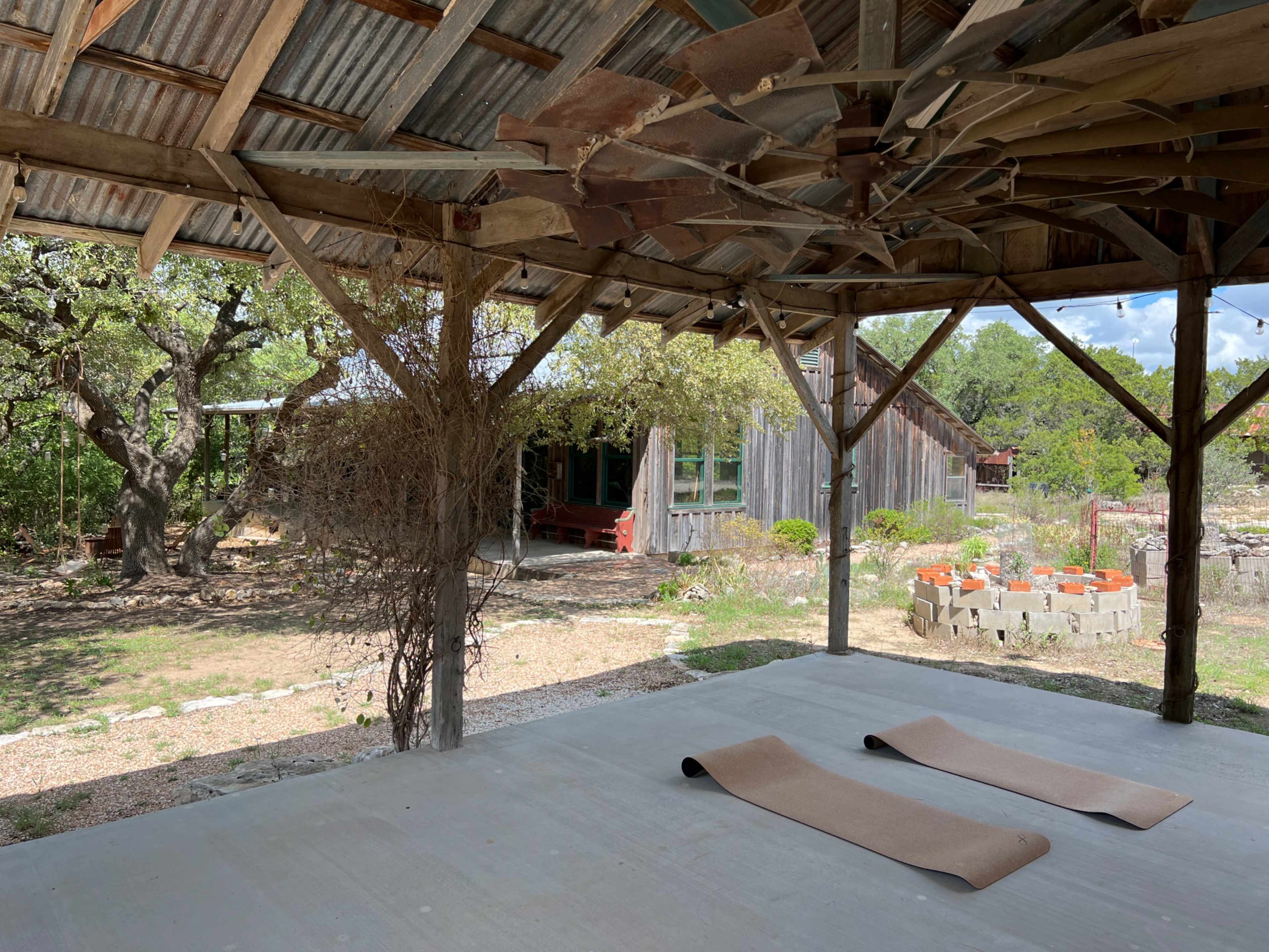 An outdoor space with two yoga mats on a concrete floor under a wooden roof, overlooking a garden area with rustic structures in the background.
