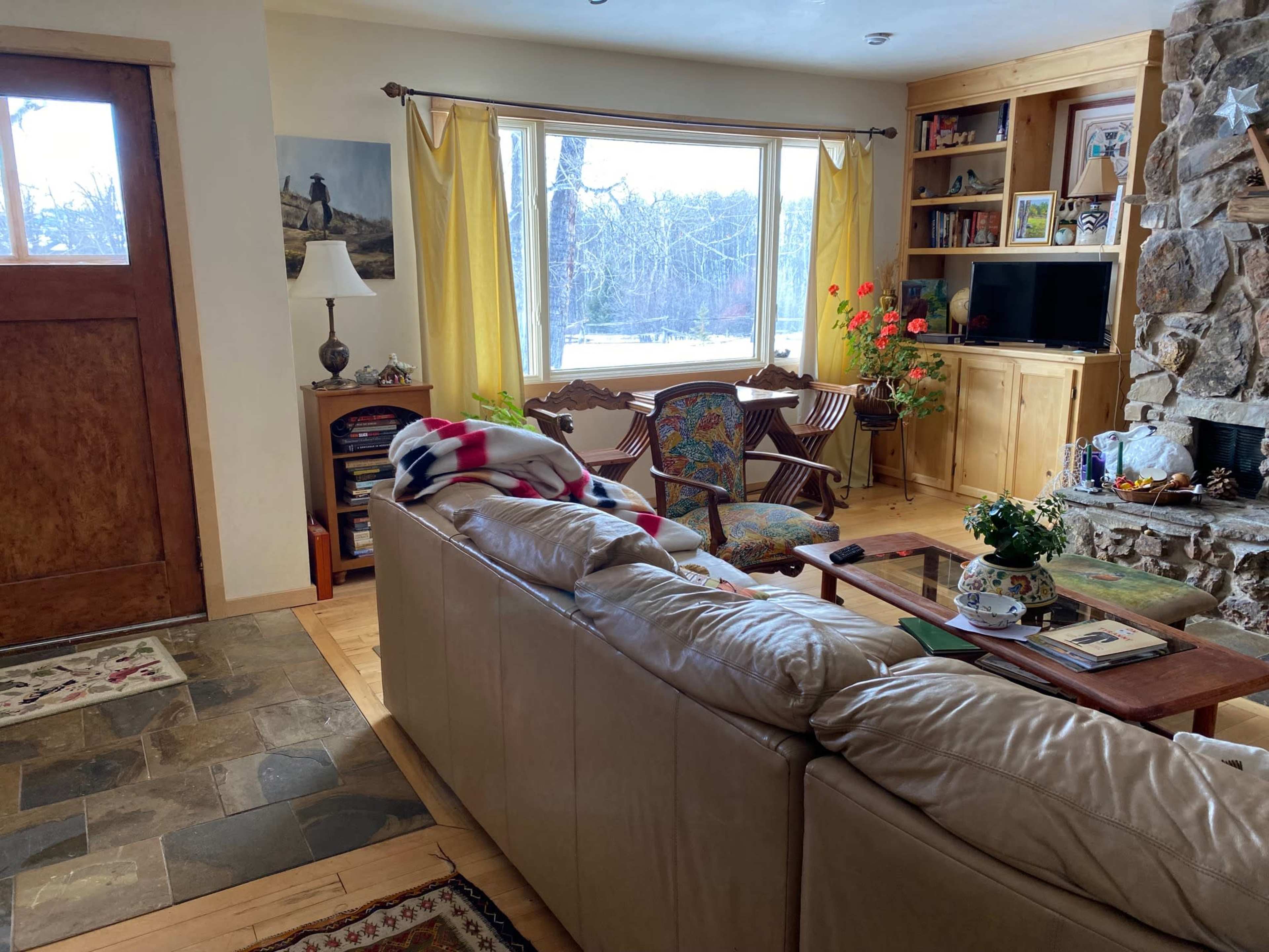 A cozy living room with a stone fireplace, a large window dressed in yellow curtains, and a mix of furniture including a leather couch and a wooden bookshelf.
