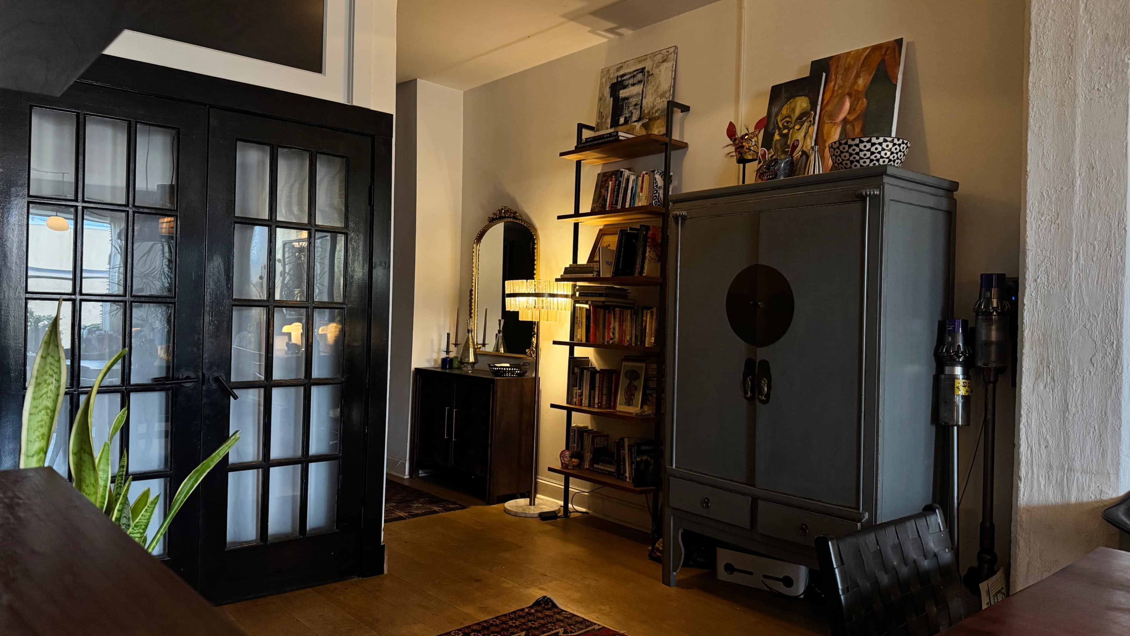 The image shows a well-furnished room featuring a vintage-style cabinet, a bookshelf with various items, and a mirror on the wall, with a doorway visible in the background.