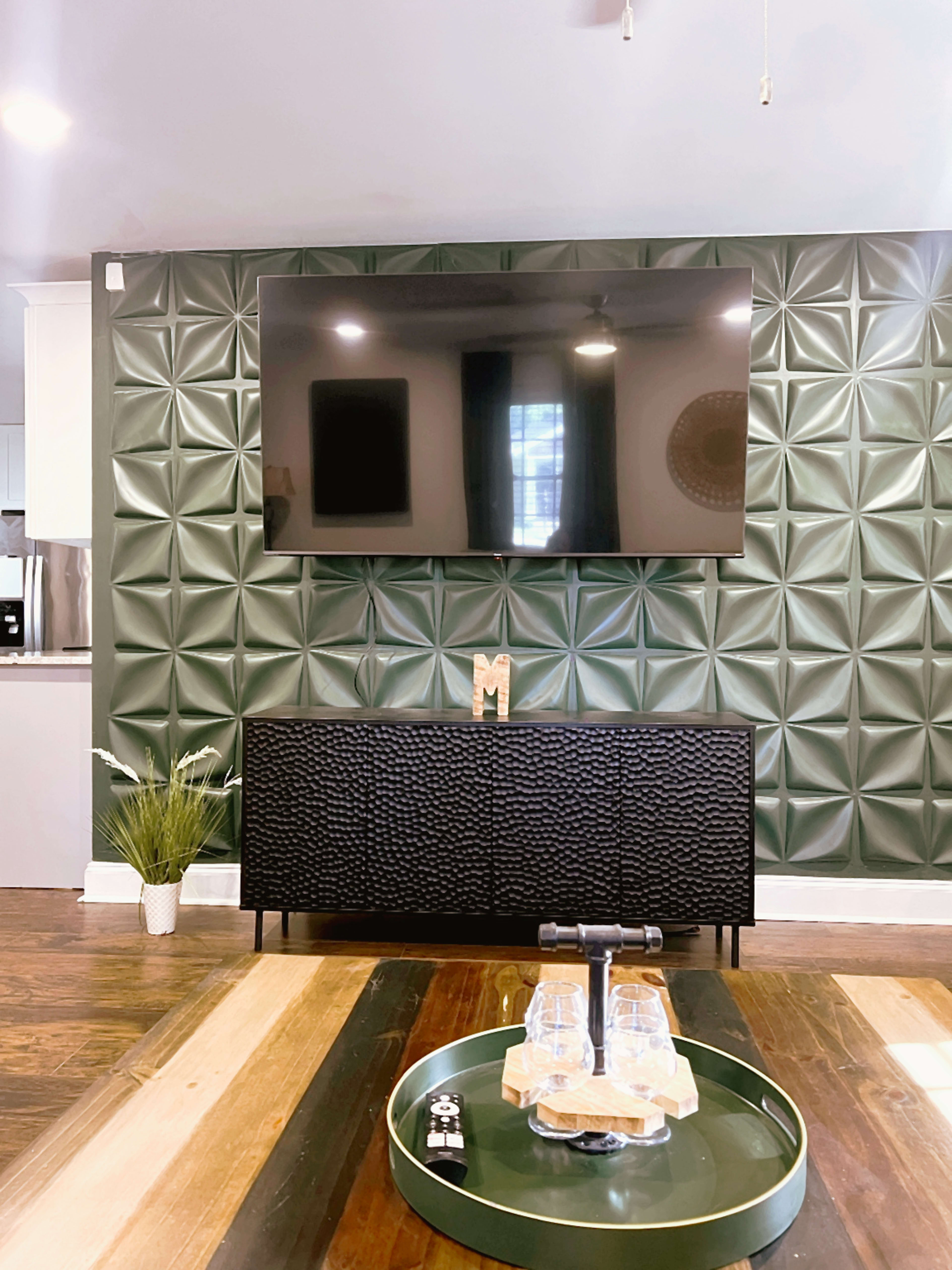 A modern living room features a textured green wall with a large flat-screen TV mounted above a black cabinet, while a coffee table displays glasses and a decorative tray.