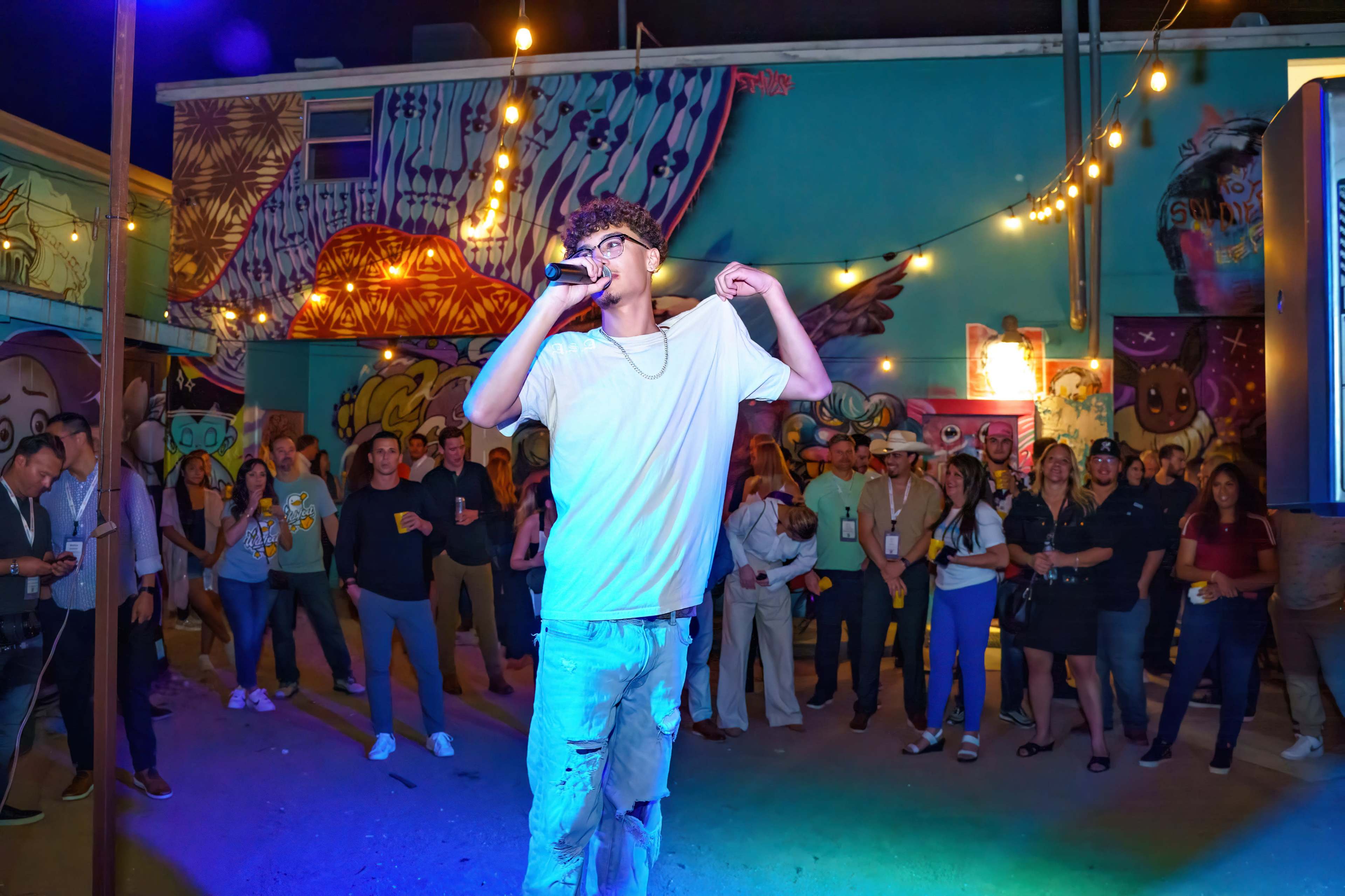 A young man performs with a microphone in front of an audience in an outdoor space adorned with colorful murals and string lights.