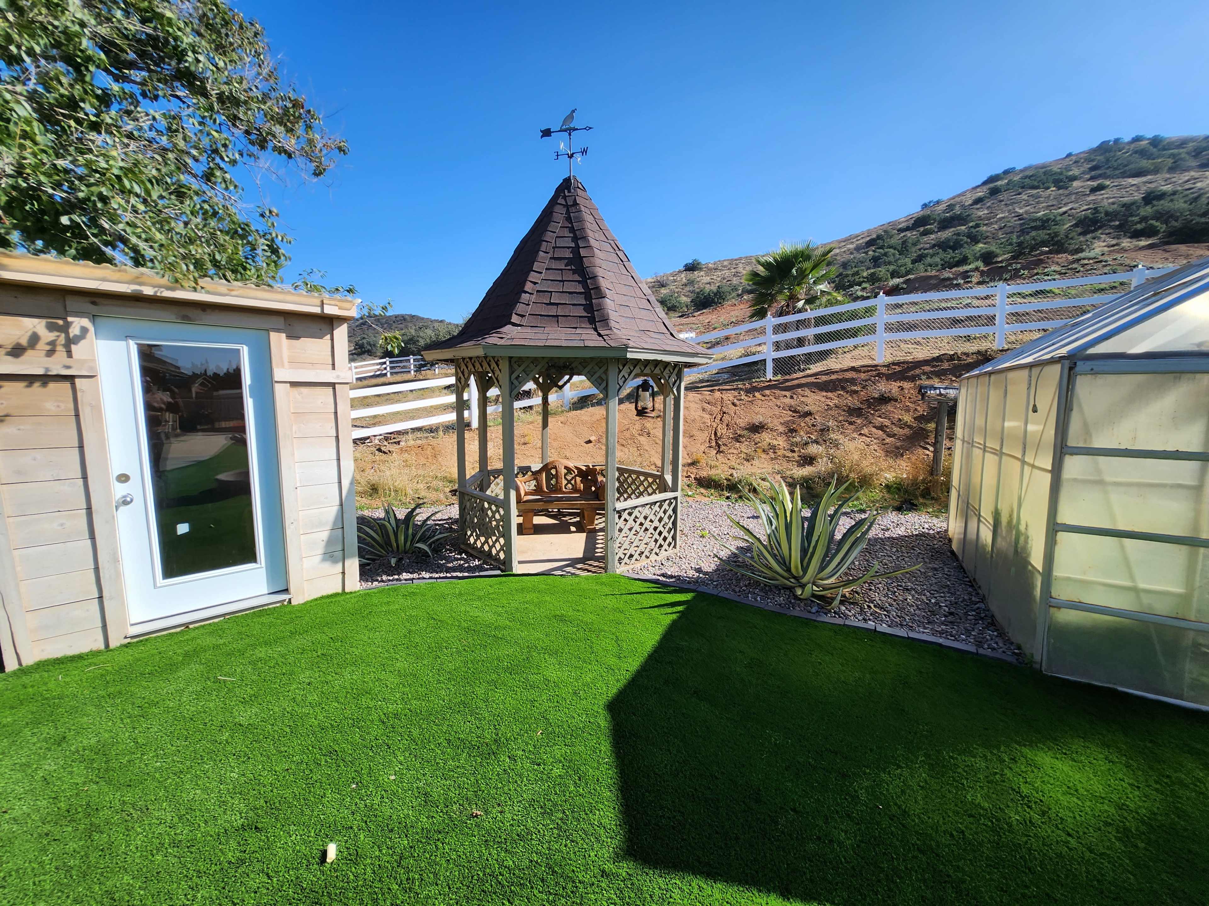 A wooden gazebo with a pointed roof is situated on a grassy area surrounded by a wooden fence and a greenhouse in a hillside garden.
