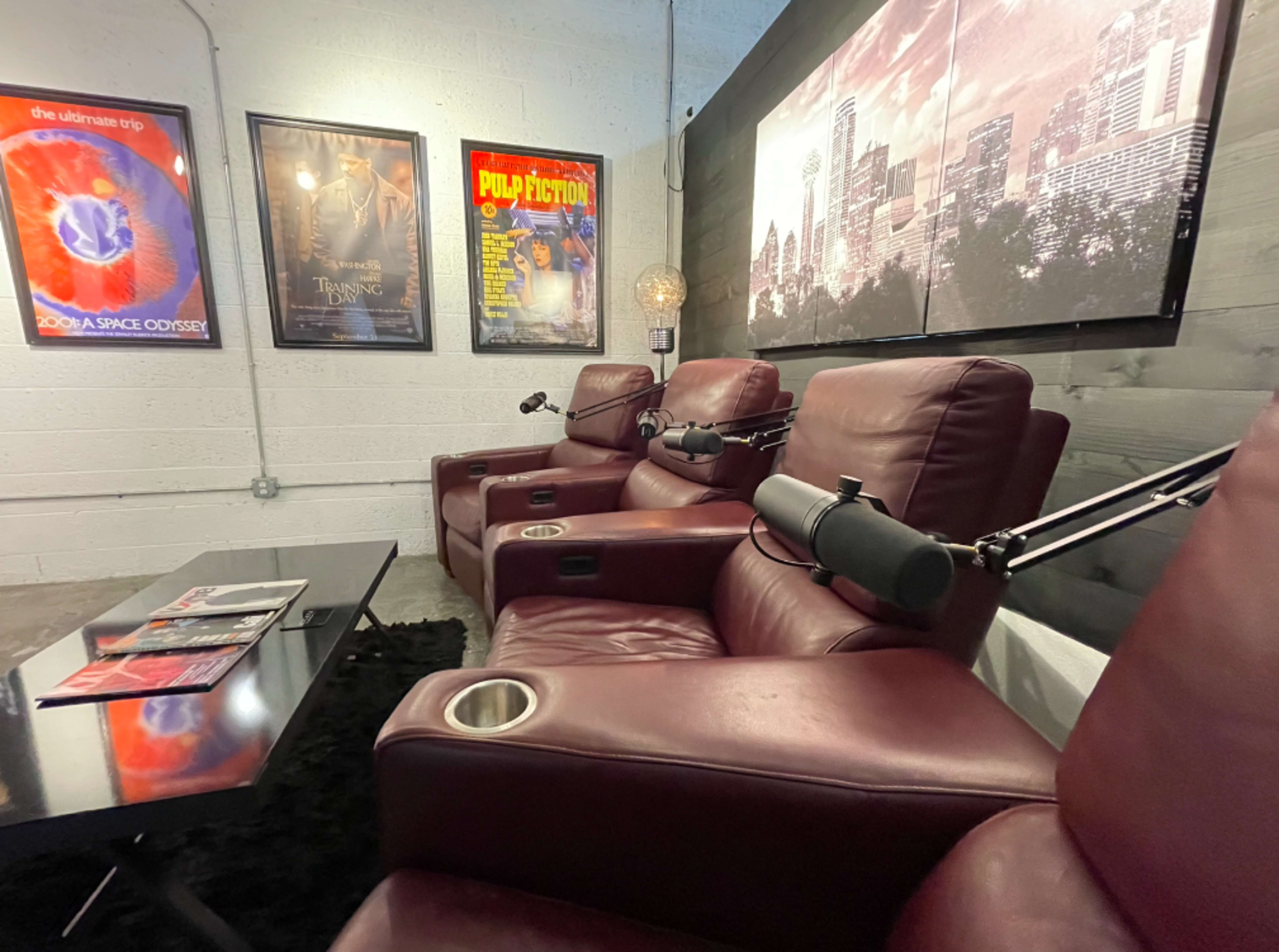 A seating area with three maroon recliners facing a wall decorated with movie posters and a small black table with magazines.
