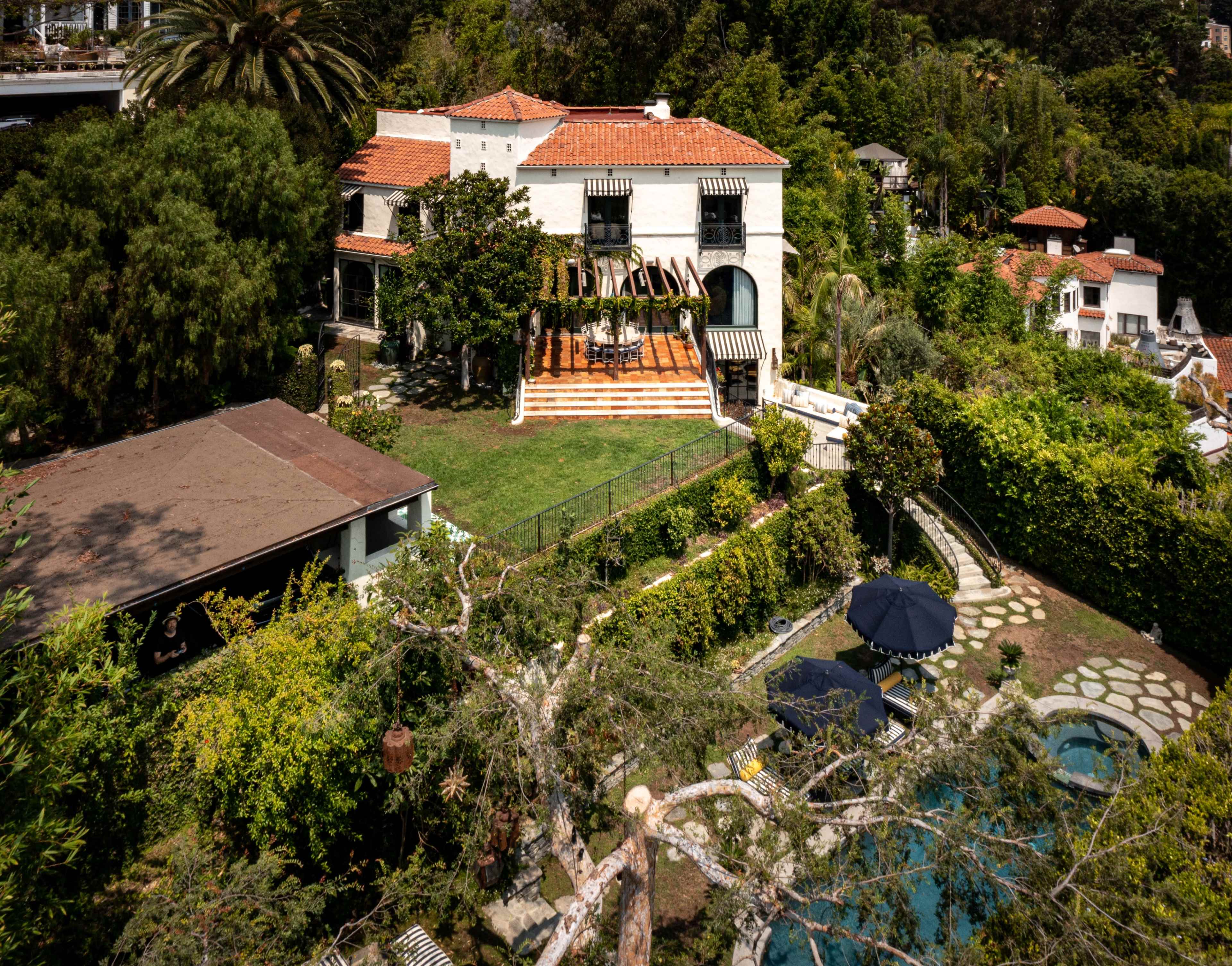 A two-story white house with a terracotta roof and a green lawn is surrounded by lush trees and a swimming pool.