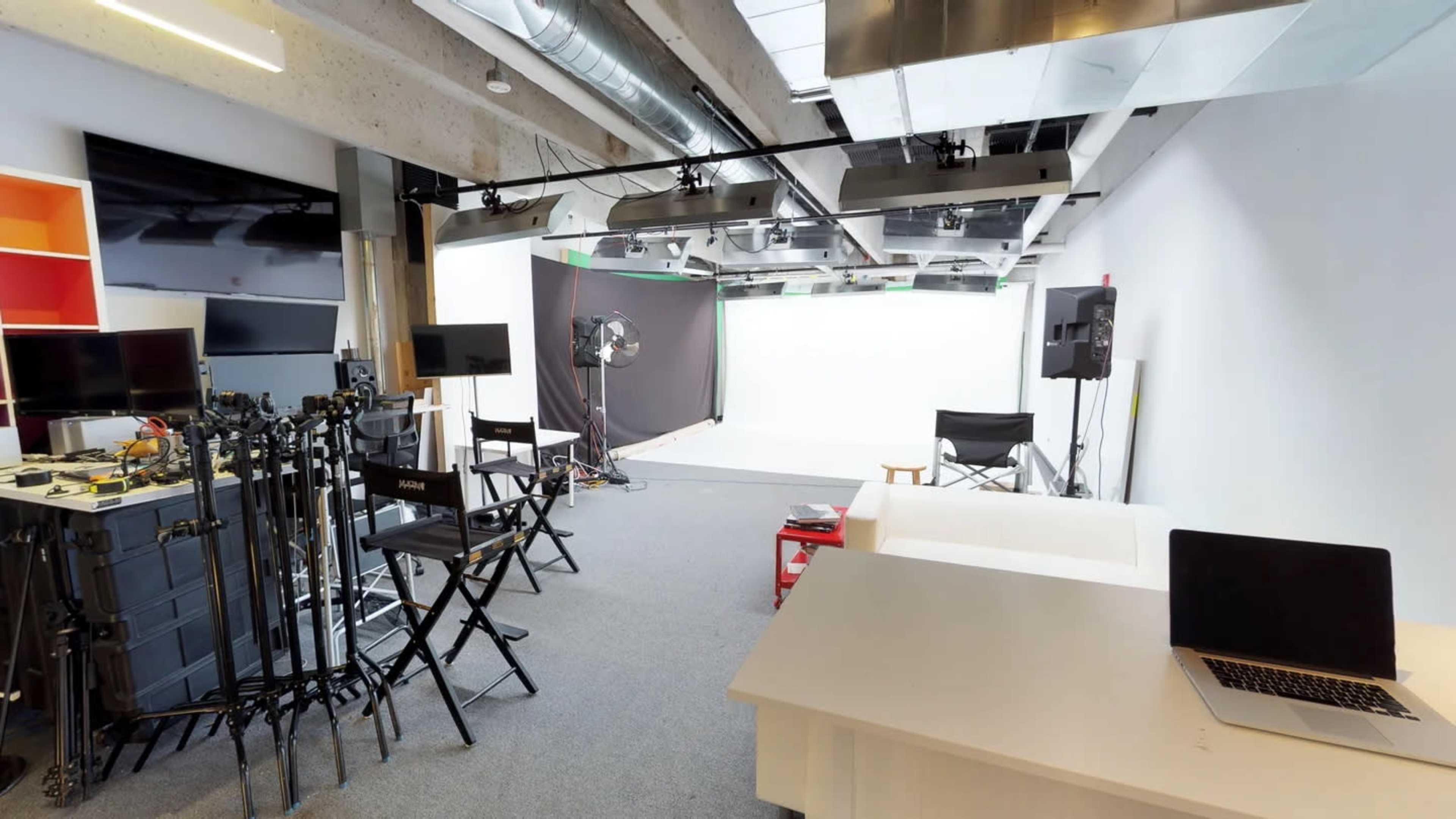The image shows a well-lit studio equipped with black chairs, camera equipment, and a white backdrop for filming.
