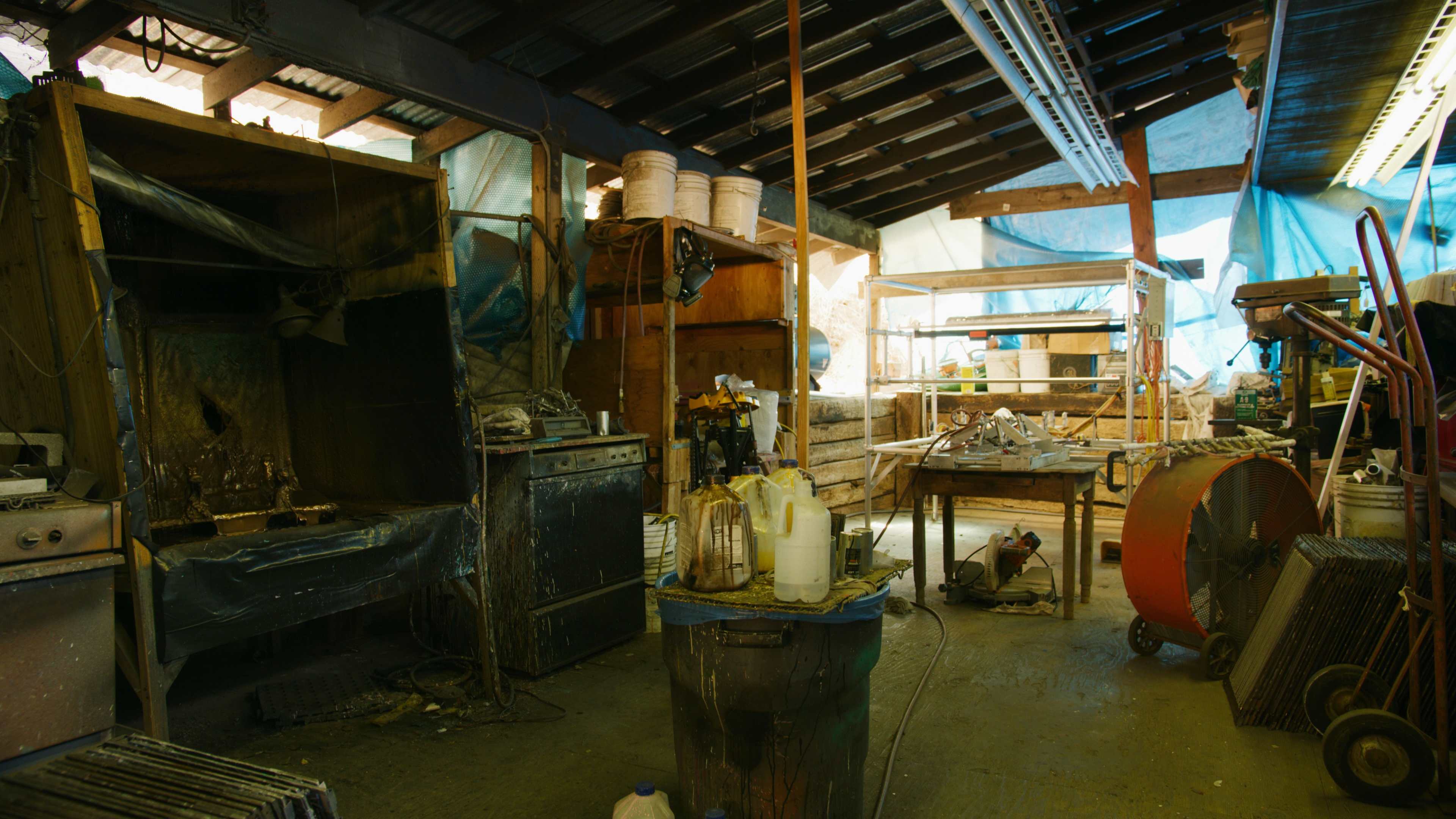 A cluttered workshop with various tools and equipment, including a stove, shelves, and buckets, under a wooden structure with a blue tarp overhead.