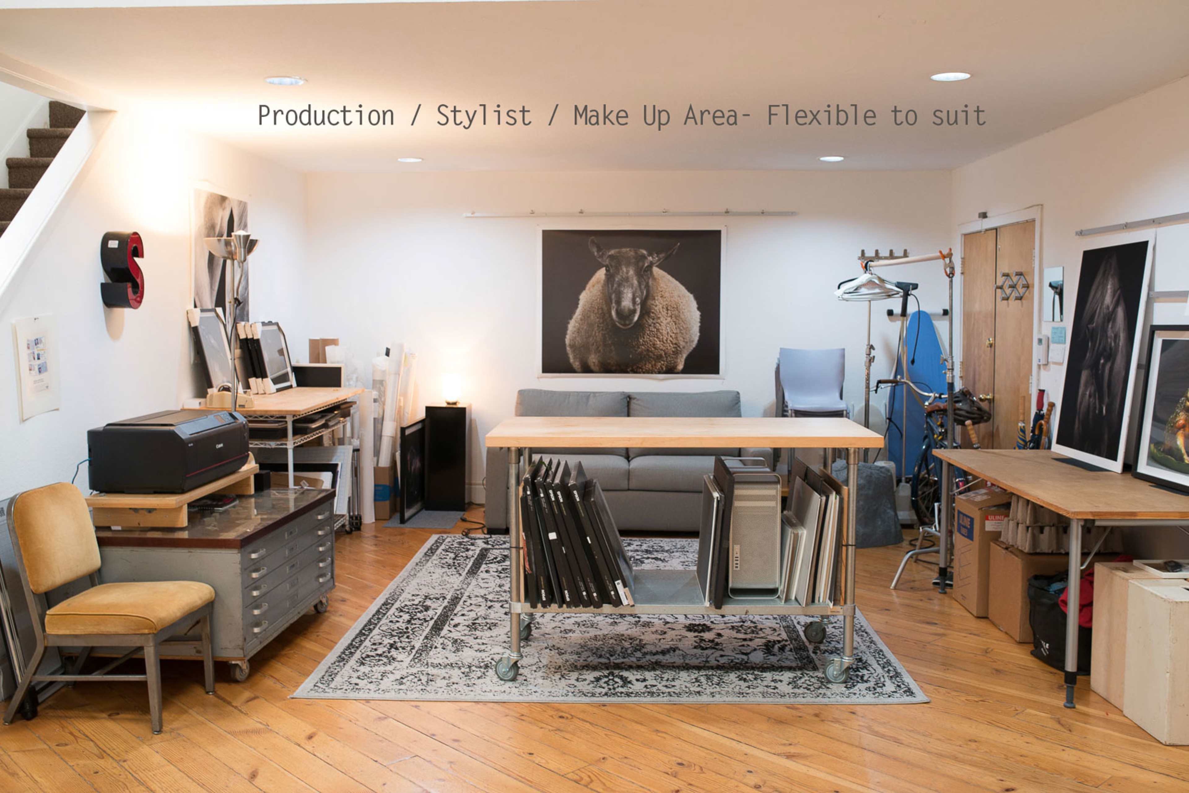 The image shows a spacious studio featuring a flexible production and styling area with a sofa, a large framed photograph of a sheep, a variety of art supplies, and equipment for styling and makeup.