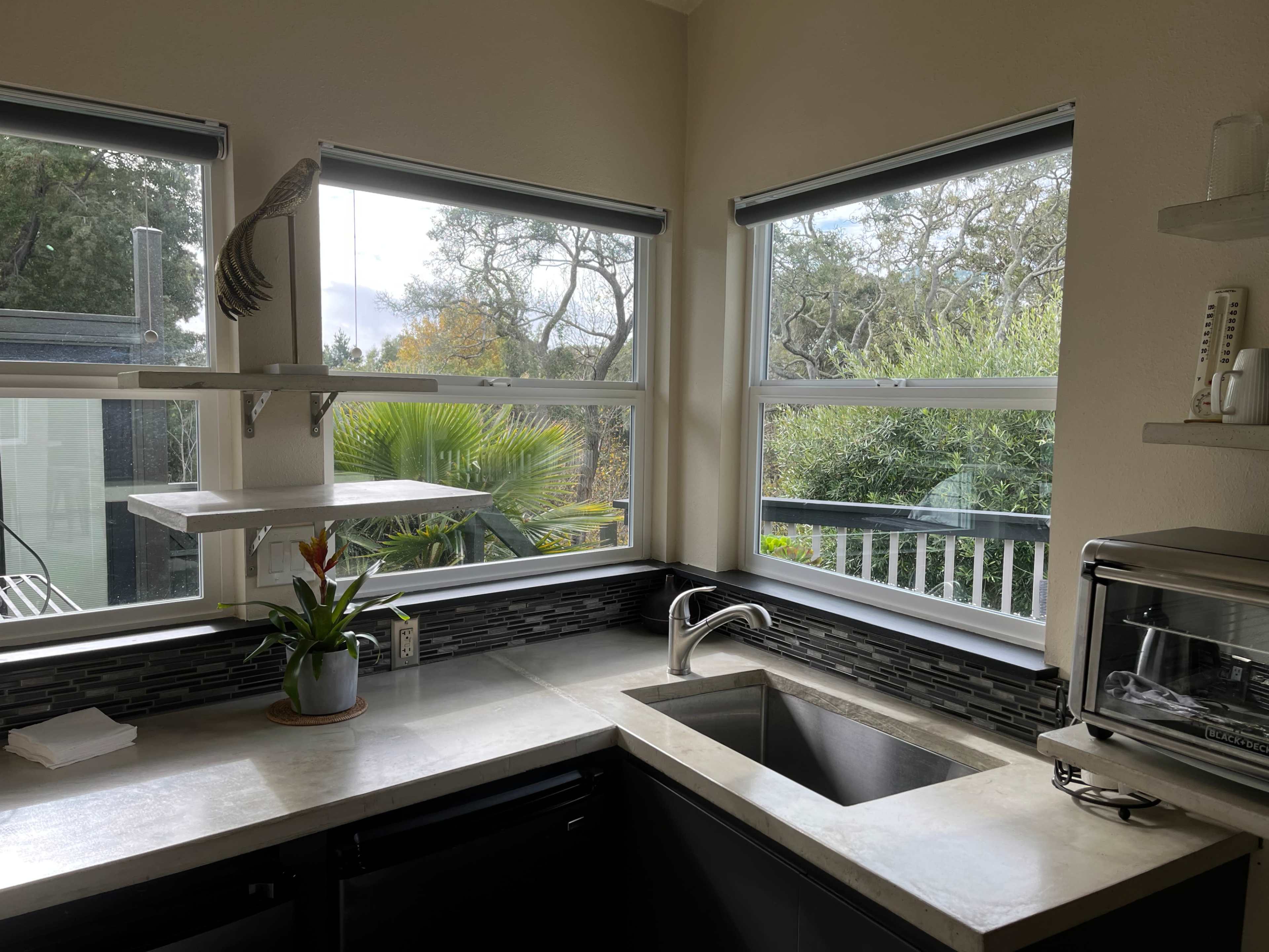 A kitchen with a stainless steel sink, a grey countertop, and large windows overlooking greenery.
