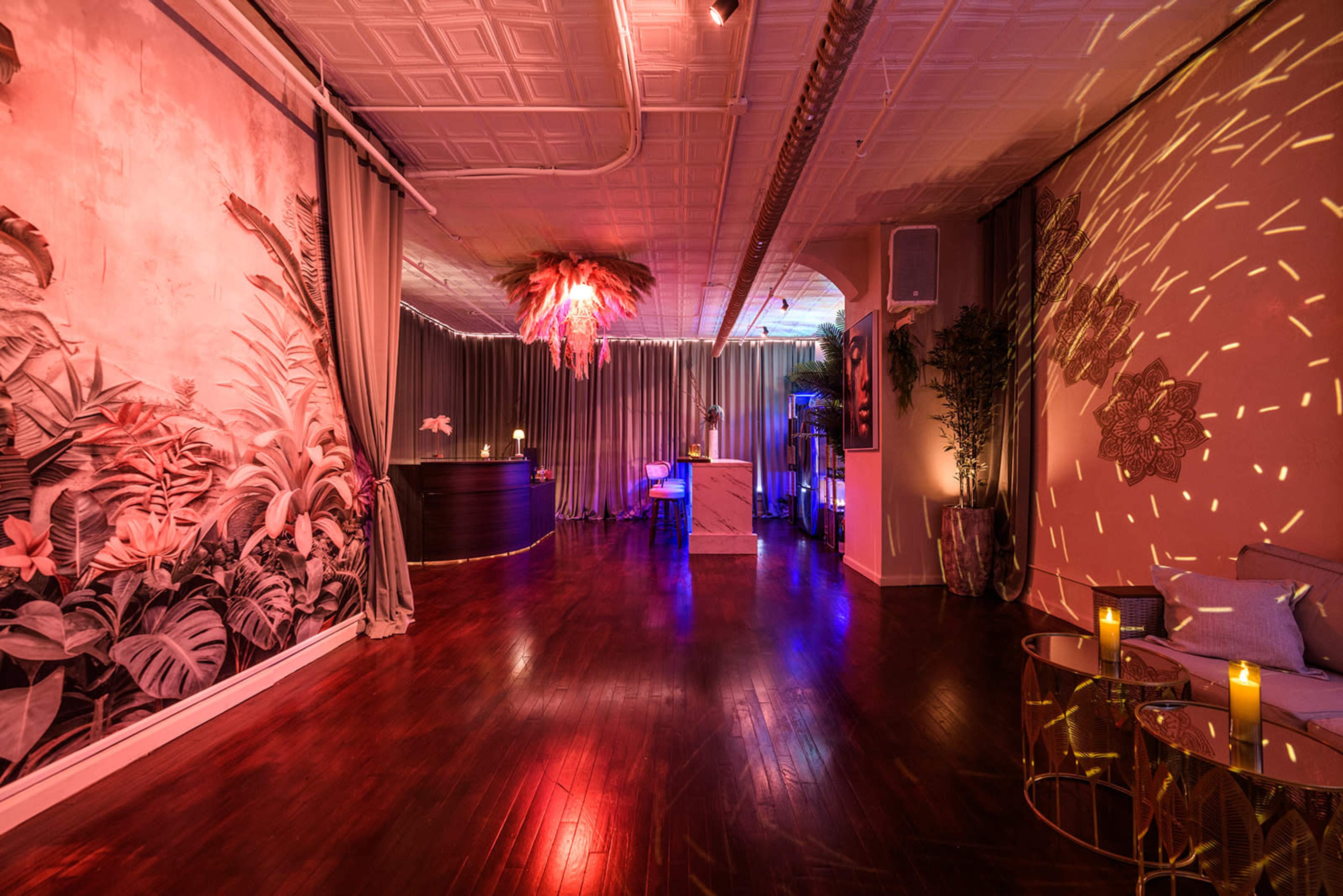 The image shows a stylish event space with a tropical-themed mural, illuminated by soft colored lighting and decorated with candles and modern furnishings.