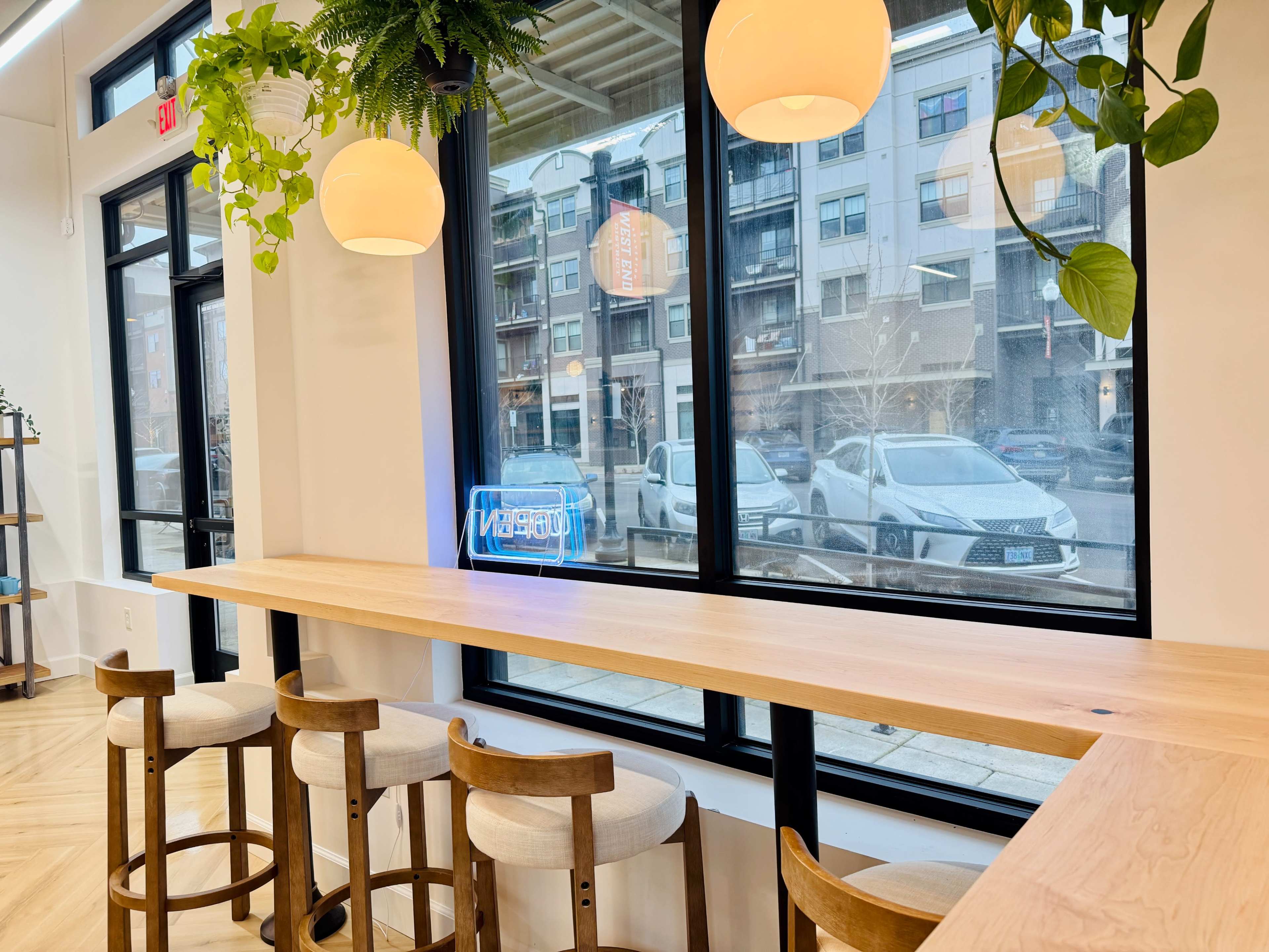A light wooden countertop with stools overlooks a large window displaying a street scene and a blue neon "OPEN" sign.