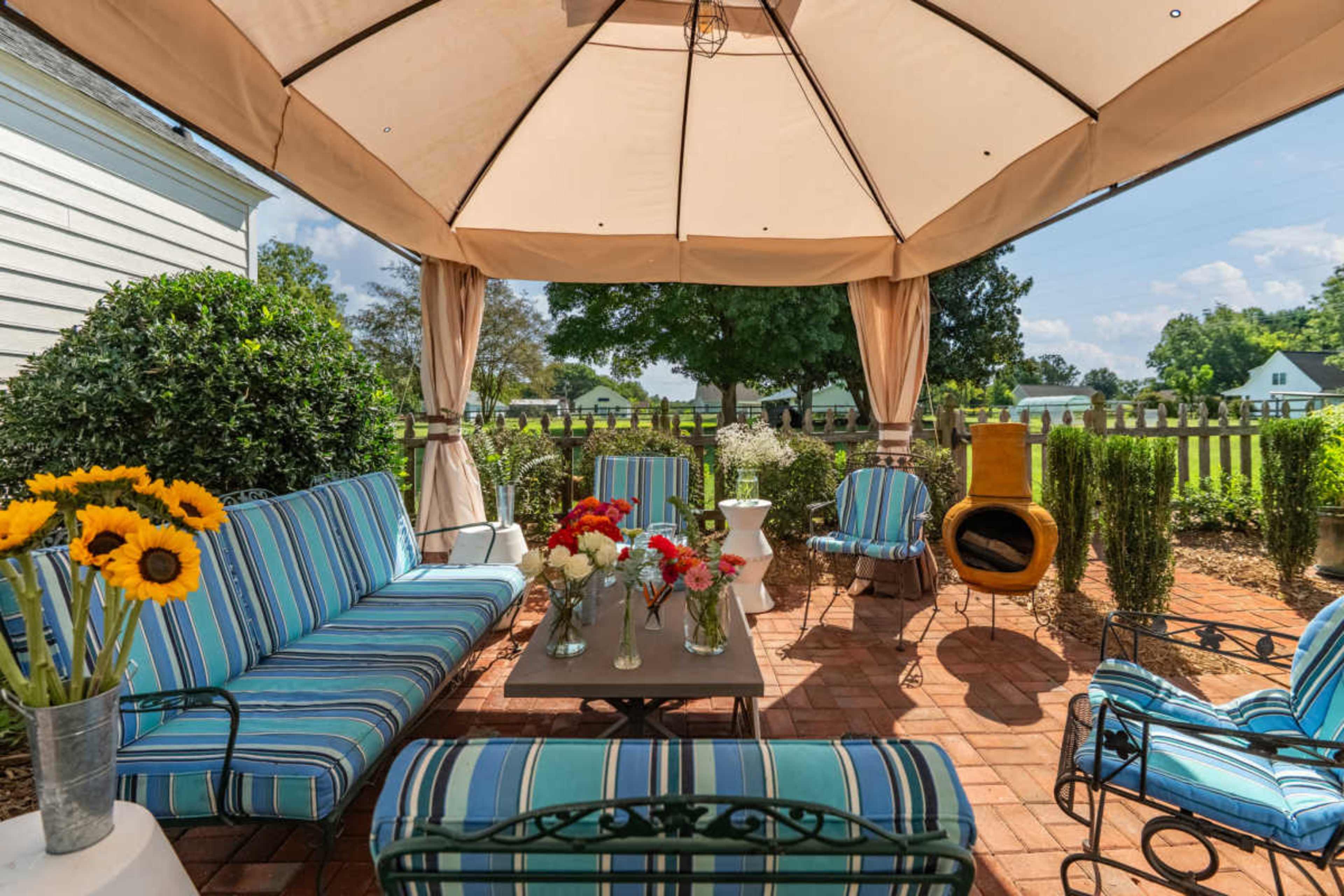 A covered outdoor seating area features striped cushions, flower arrangements, and a brick patio surrounded by greenery.