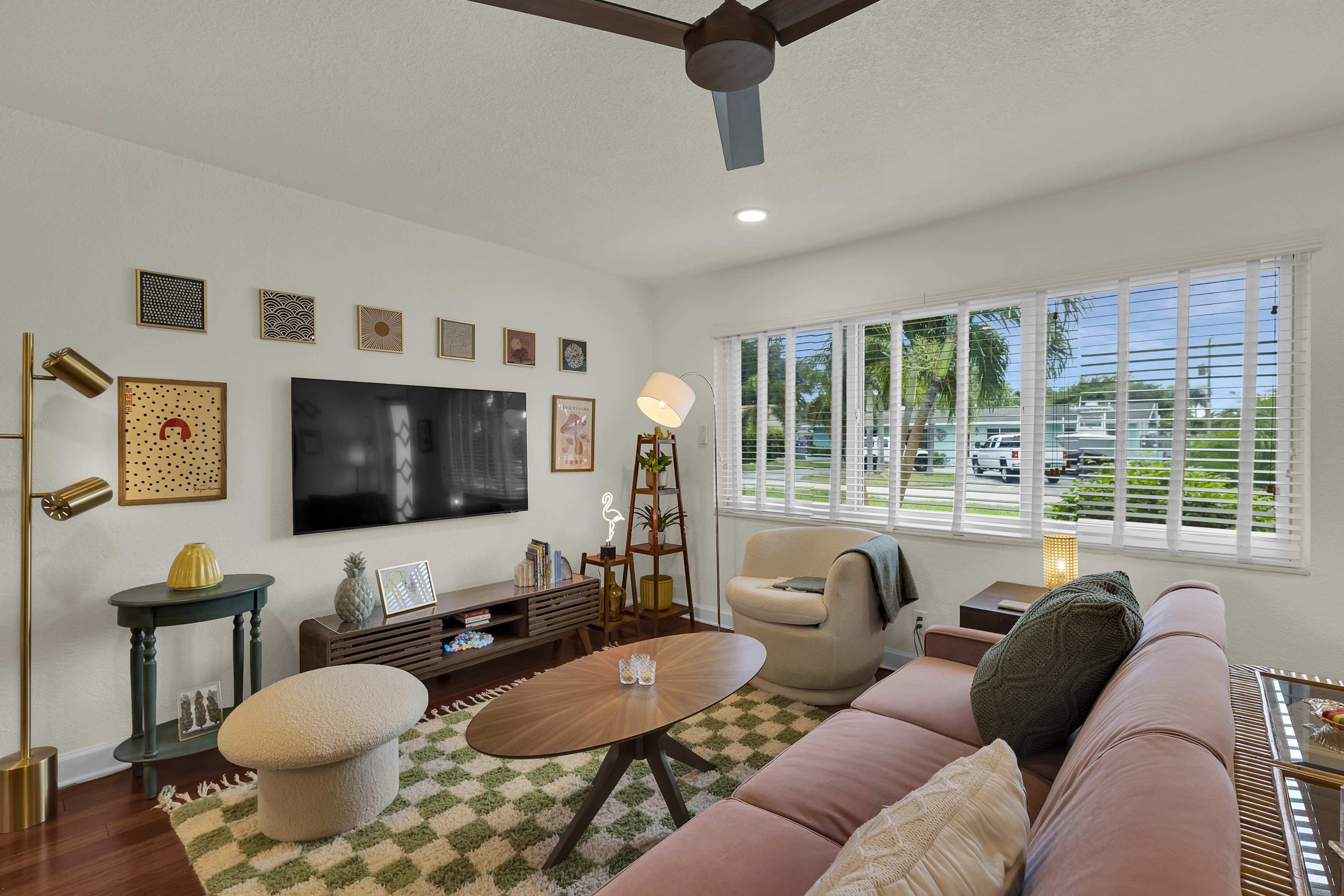 The living room features a pink sofa, a wooden coffee table, and a large television mounted on the wall, with windows allowing natural light to illuminate the space.
