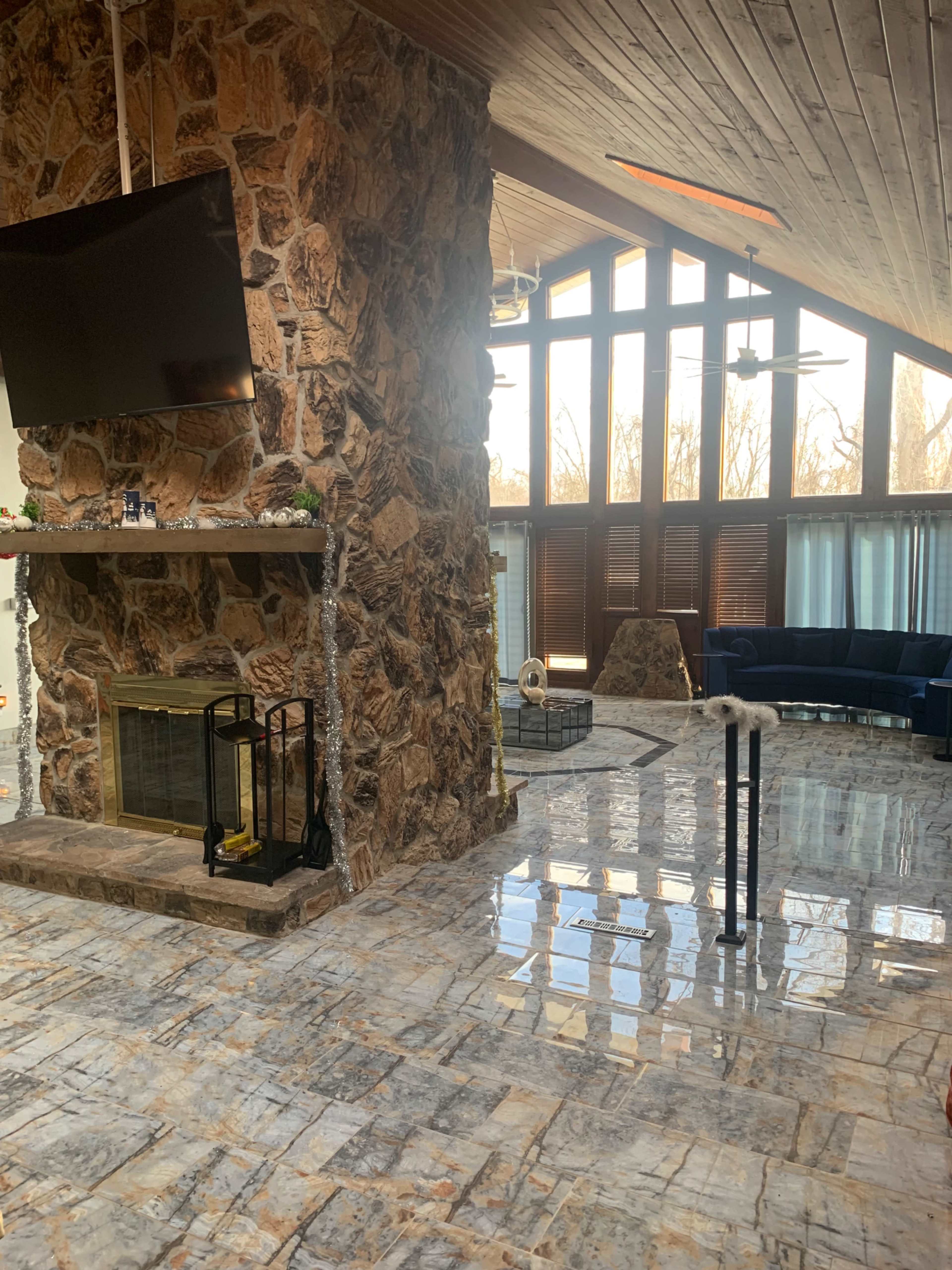 The image shows a spacious living room featuring a stone fireplace, a large television mounted on the wall, and large windows that allow natural light to fill the space.