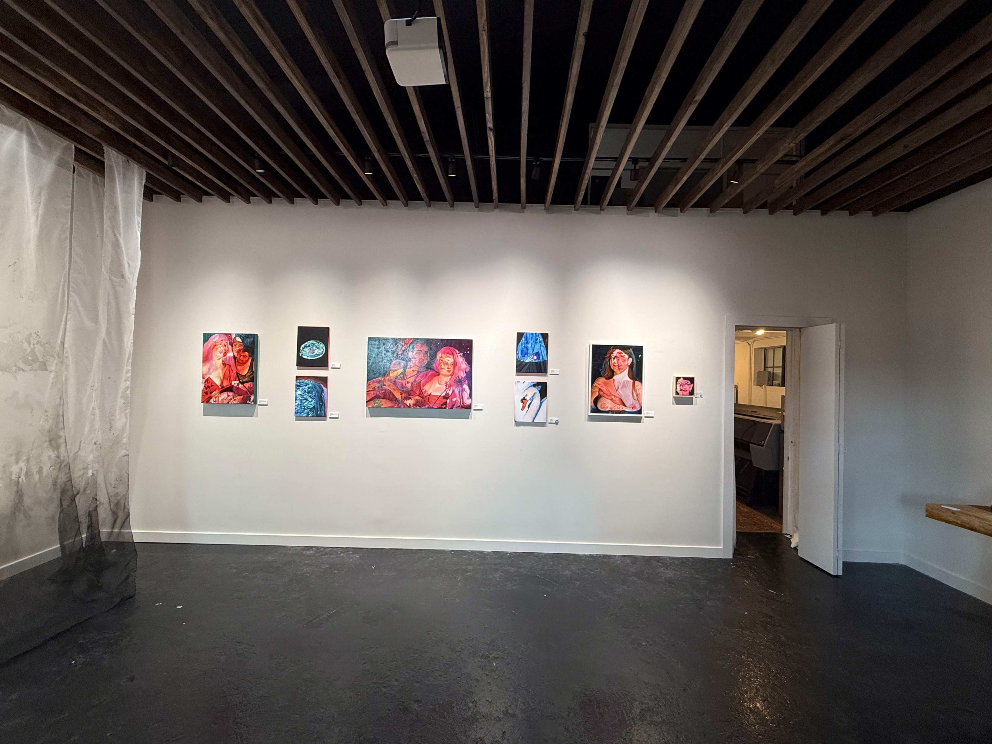 The image shows an art gallery interior featuring a wall with several colorful artworks displayed, alongside a partially visible doorway leading to another room.