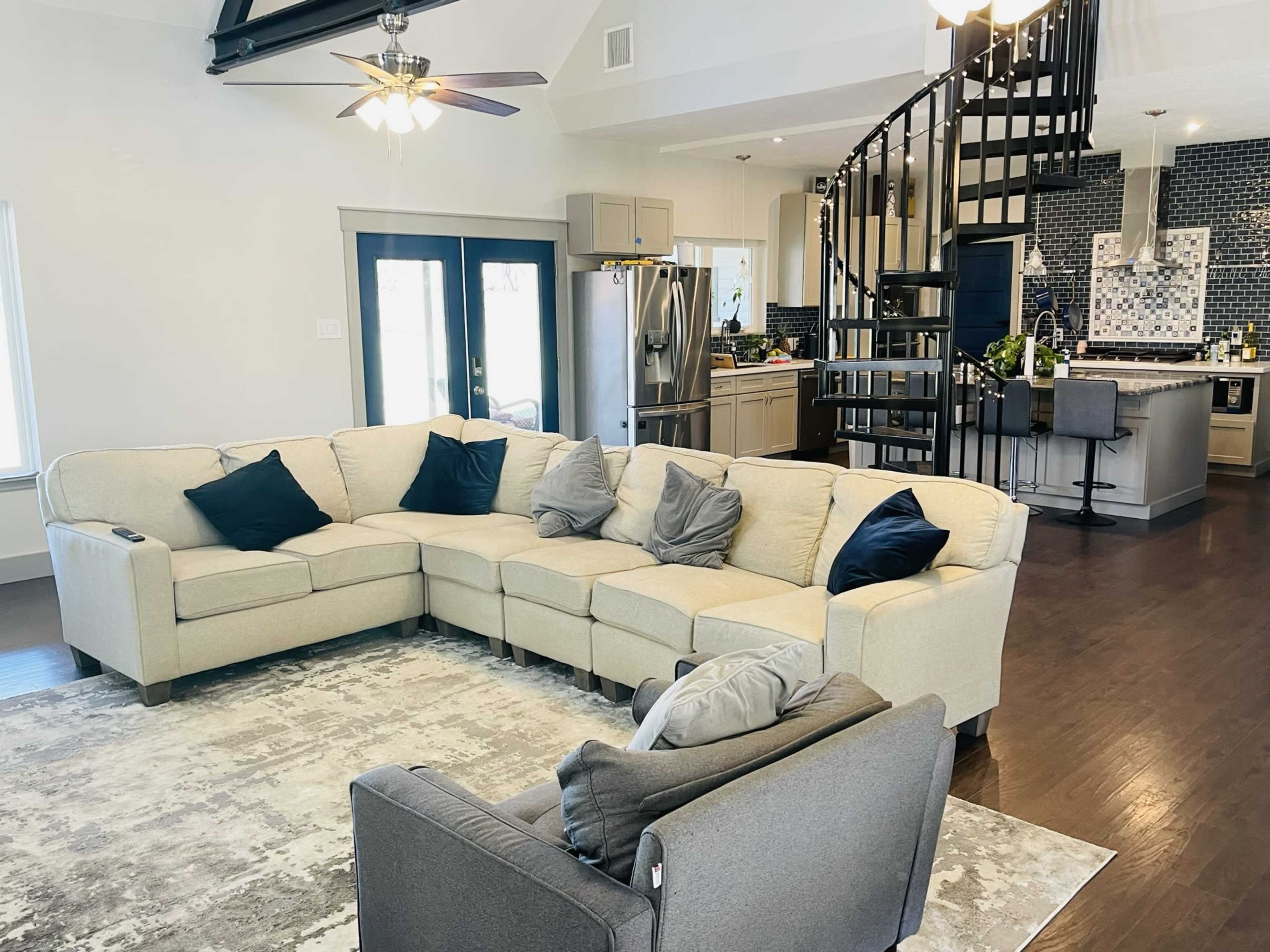 A spacious living area features a large sectional sofa with dark pillows, overlooking a modern kitchen with a spiral staircase in the background.