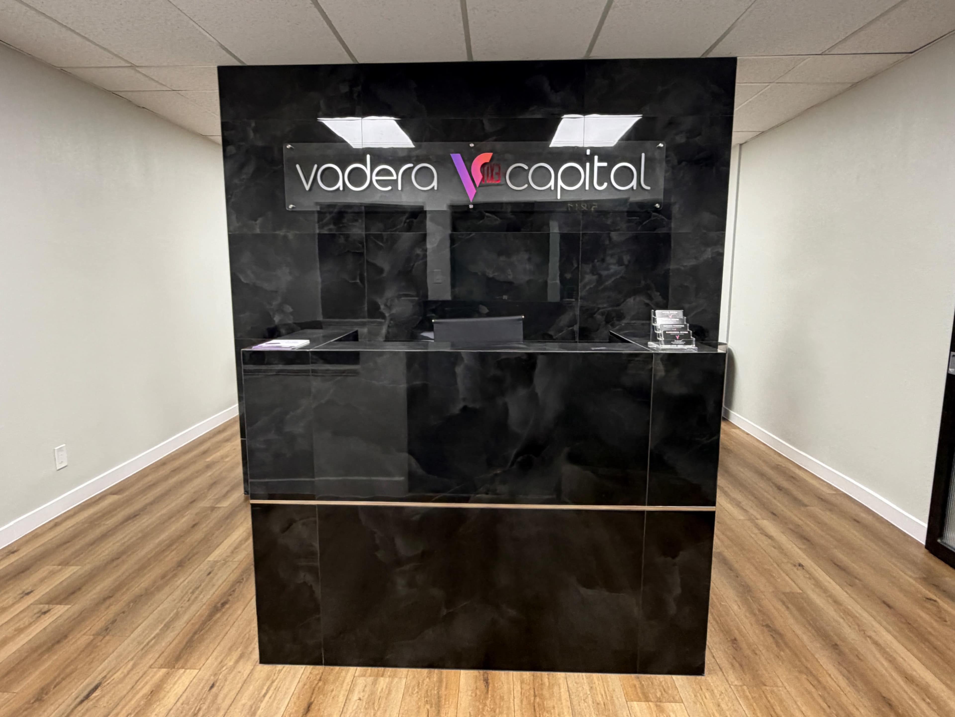 The image shows a modern reception desk with a glossy black surface and a sign that reads "vadera capital."