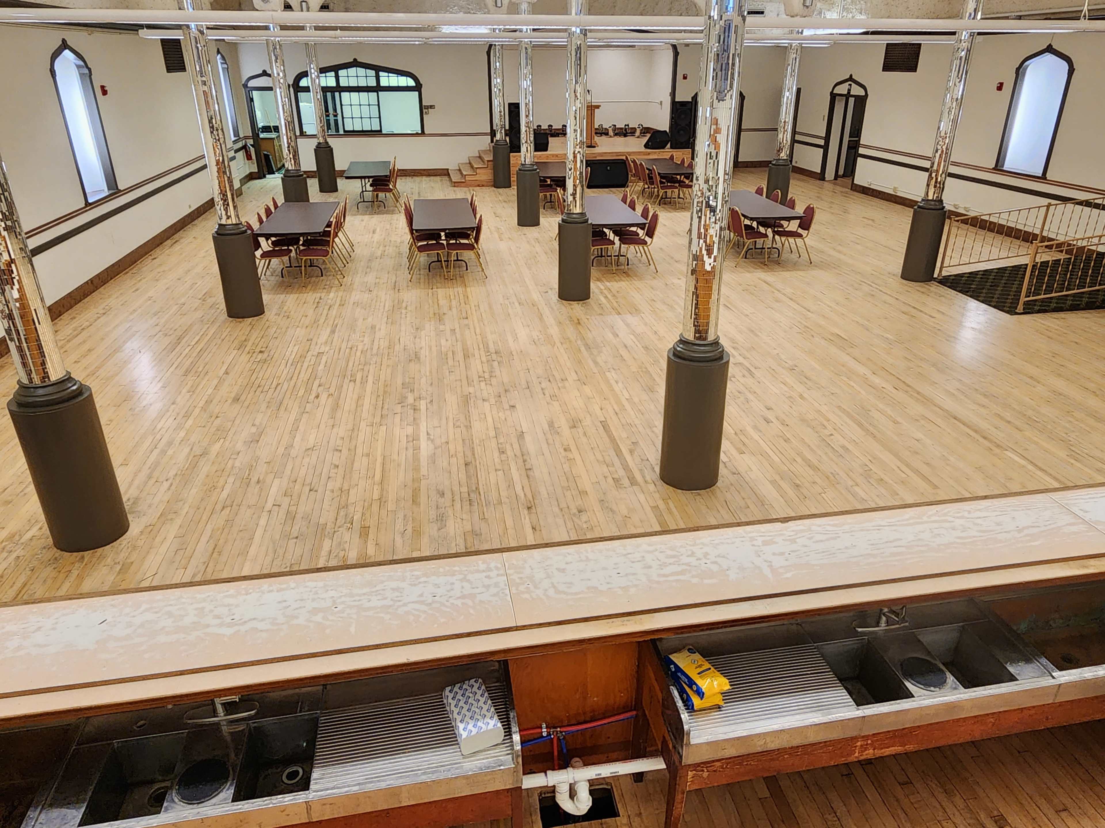 The image shows a large, empty event space with polished wooden floors, rows of tables and chairs, and a bar area with sinks in the foreground.