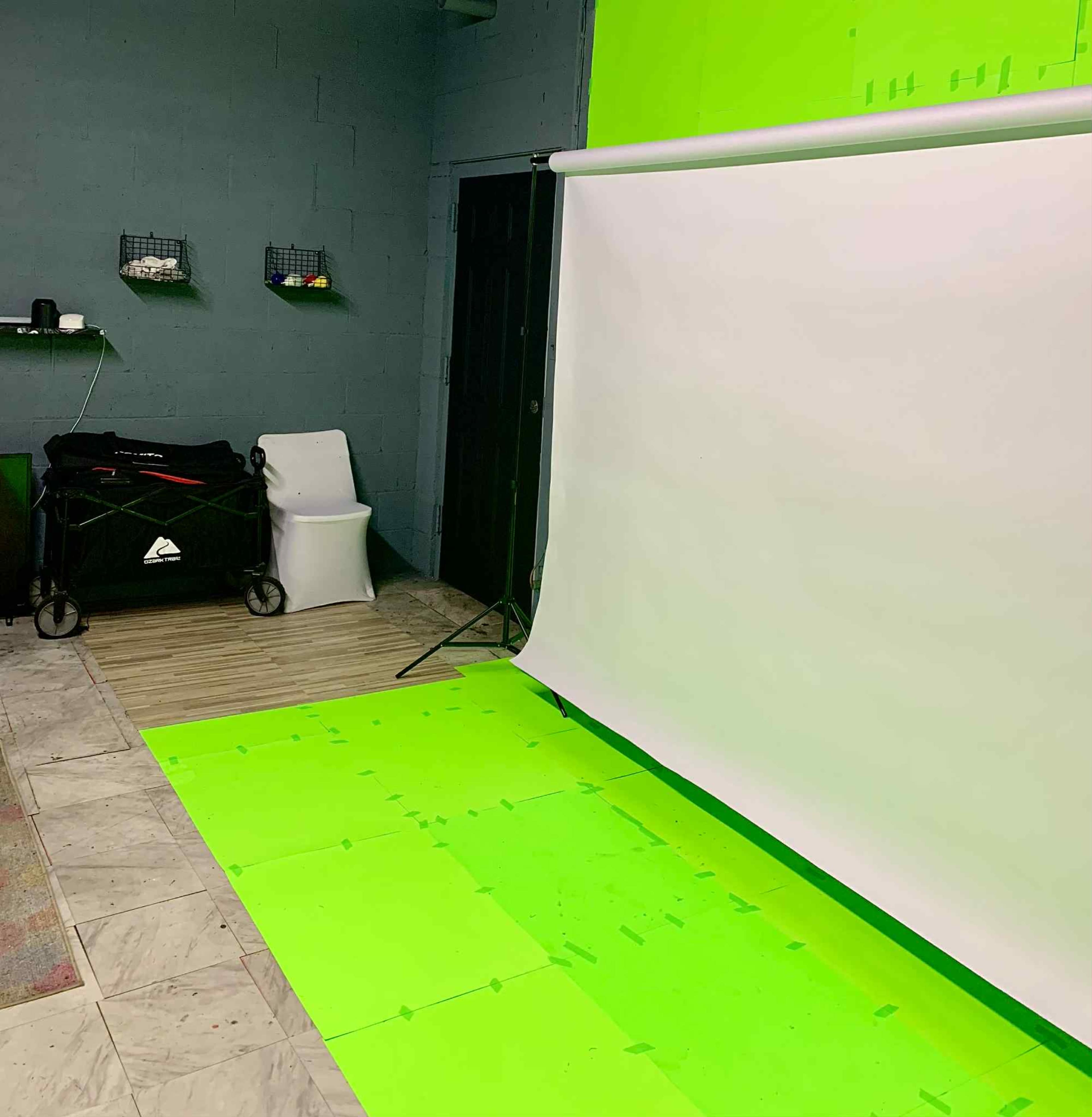 The image shows a studio setup featuring a green backdrop and a white screen, with a restroom and equipment cart visible in the background.