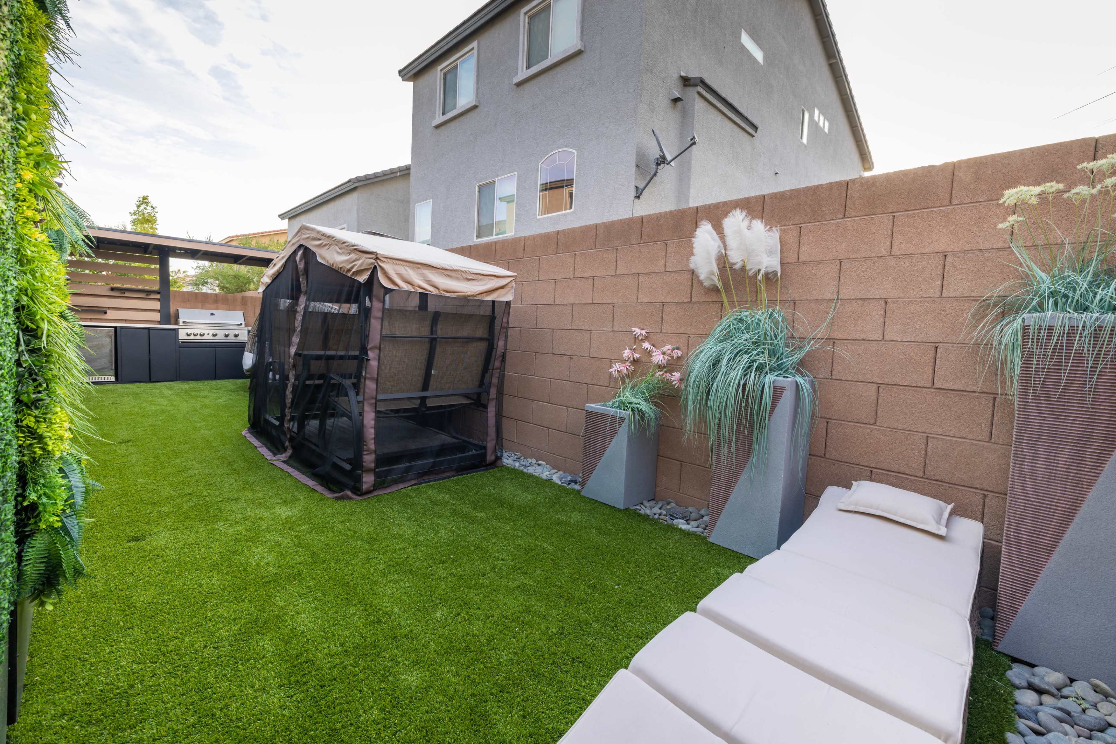 The image shows a small outdoor area with artificial grass, a patio structure covered with a tarp, and planters with decorative grass.