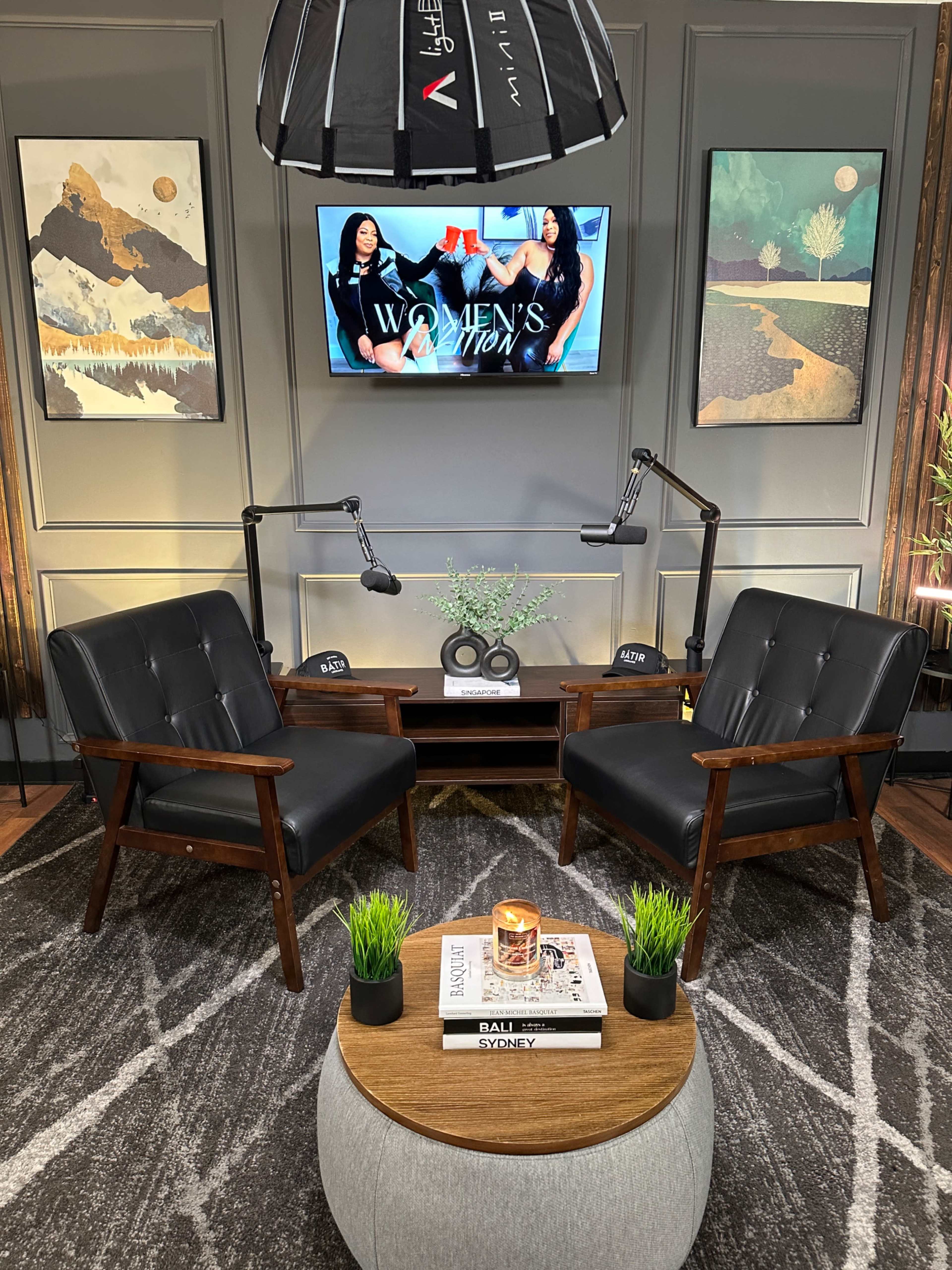 The image shows a well-decorated room featuring two black armchairs facing a coffee table, with a television displaying a women's show and artwork on the walls.