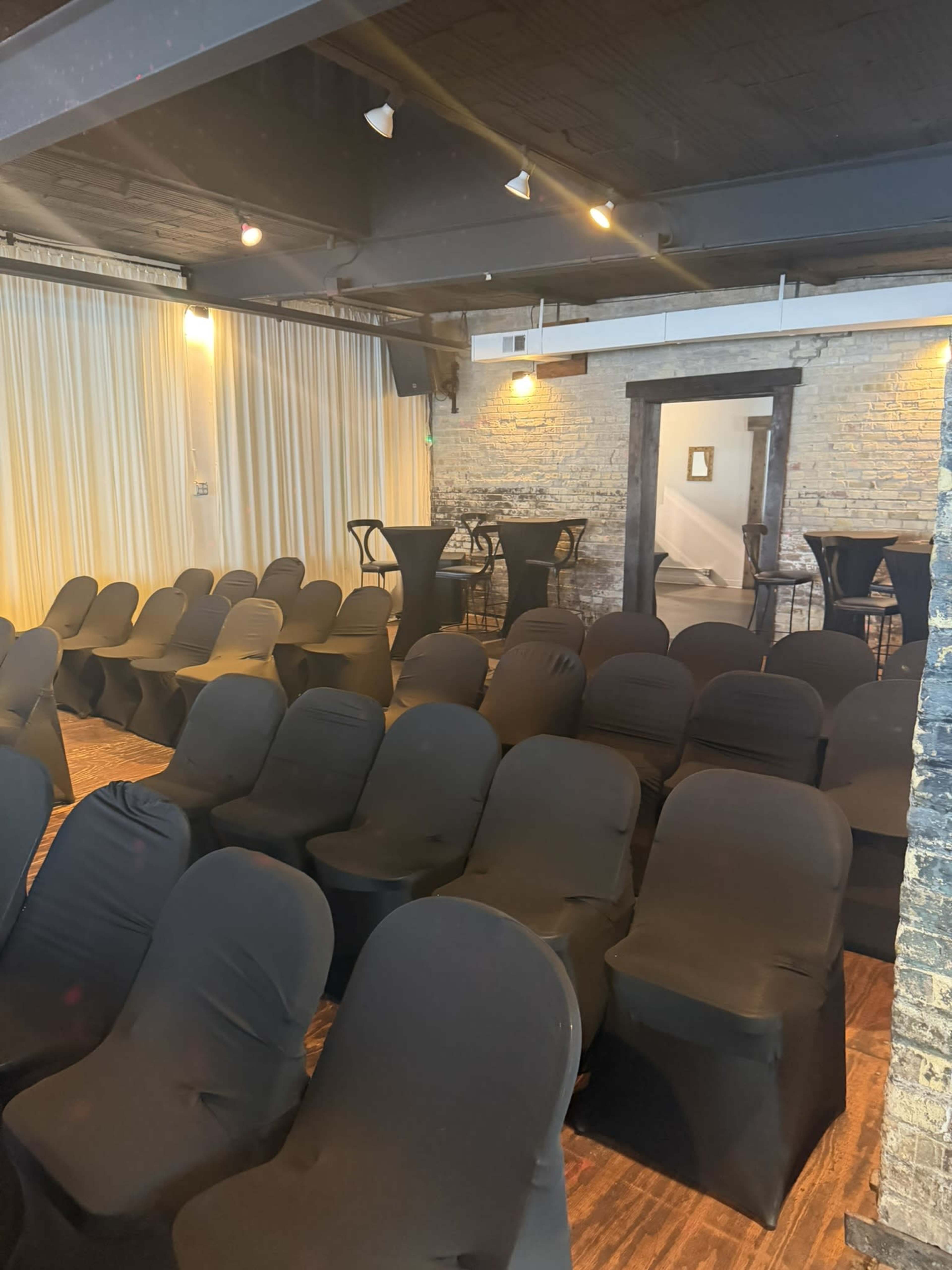 The image shows a seating arrangement of black chair covers set up in a venue with exposed brick walls and dim lighting.