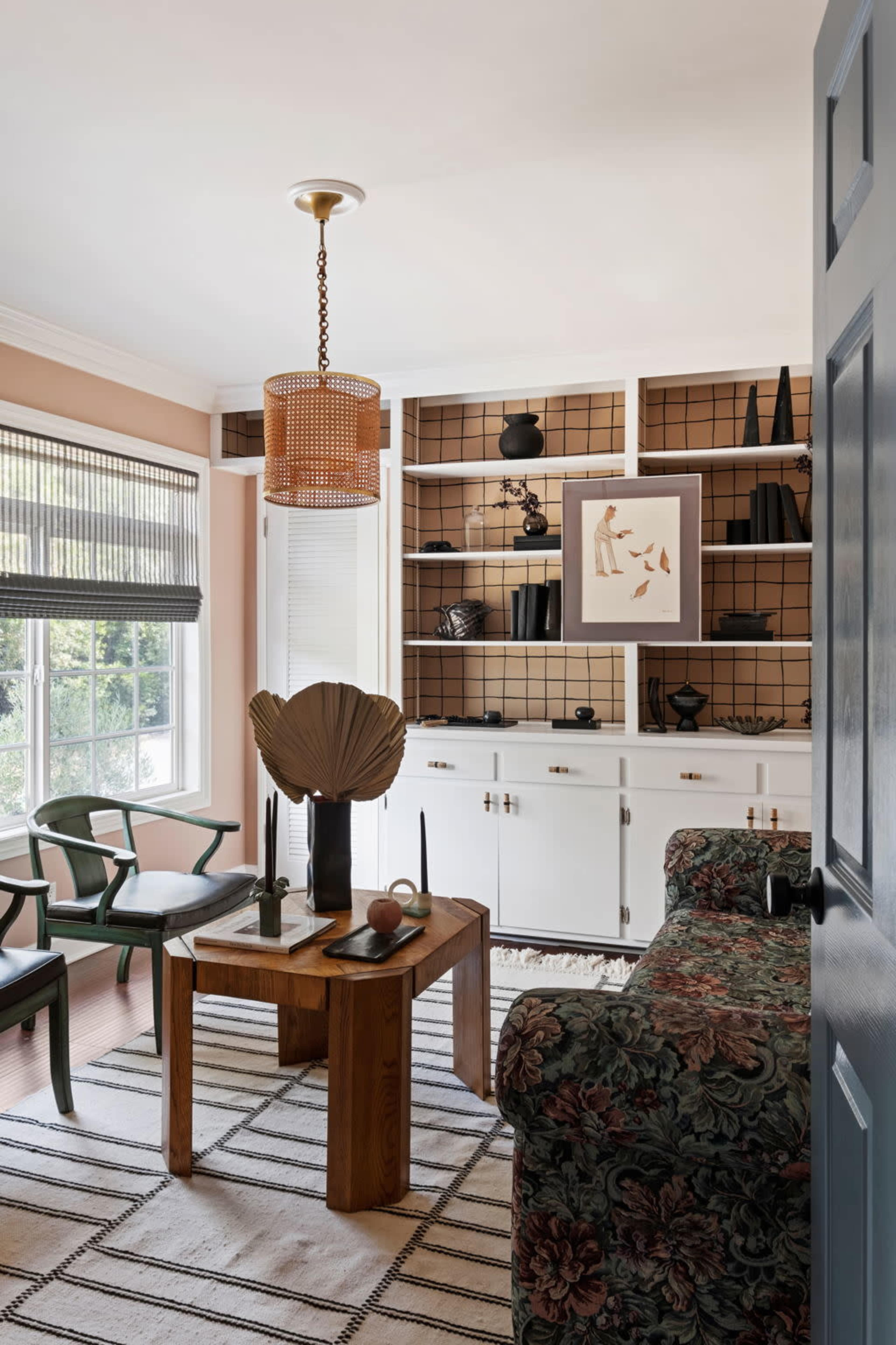 A cozy living room features a floral-patterned sofa, a wooden coffee table, and a shelf with various decorative items against a light-colored wall.