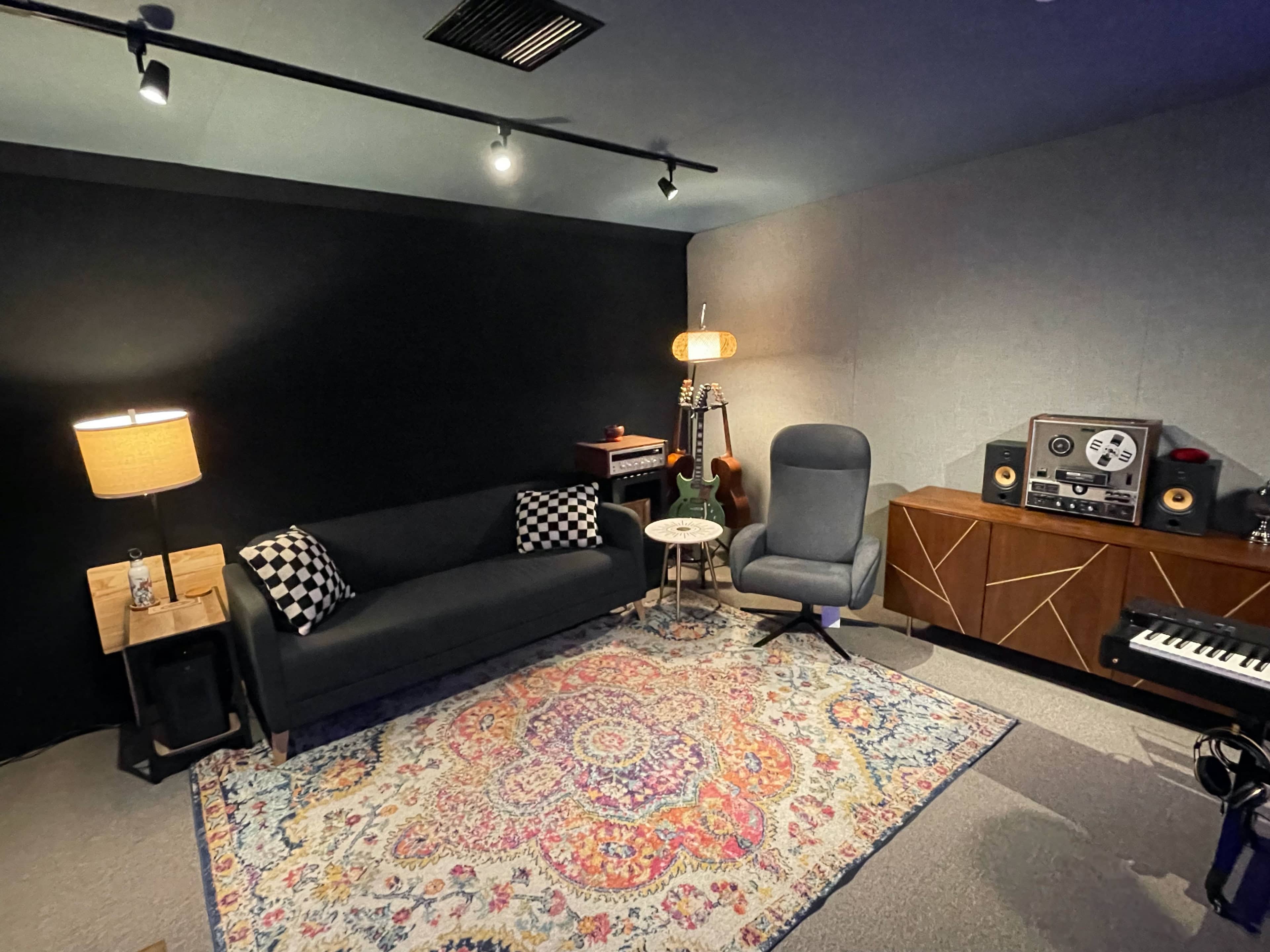 A cozy studio room featuring a black sofa, a patterned rug, a small table, a lamp, musical instruments, and audio equipment.