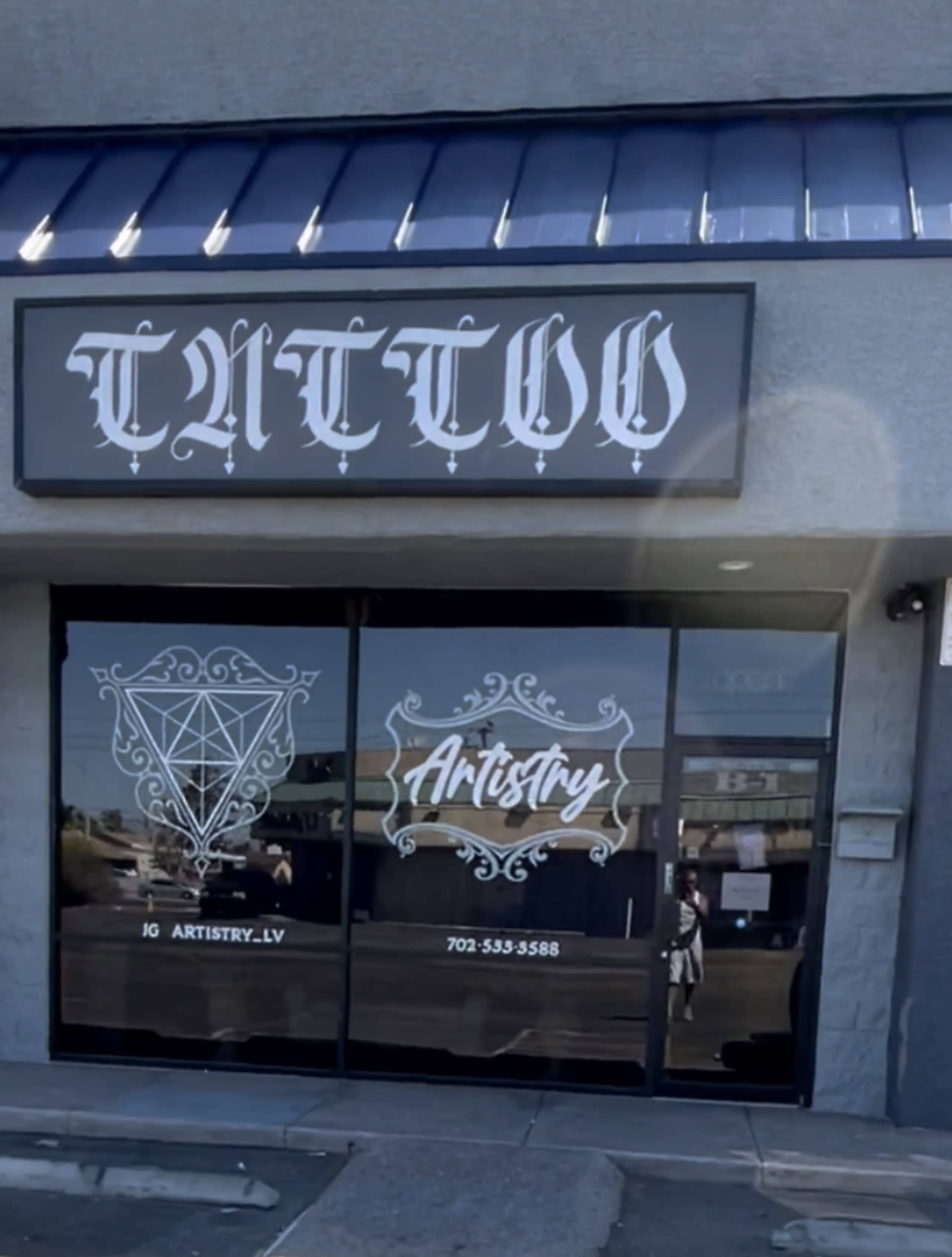 The storefront of a tattoo shop named "Artistry," featuring large windows with decorative designs and contact information.