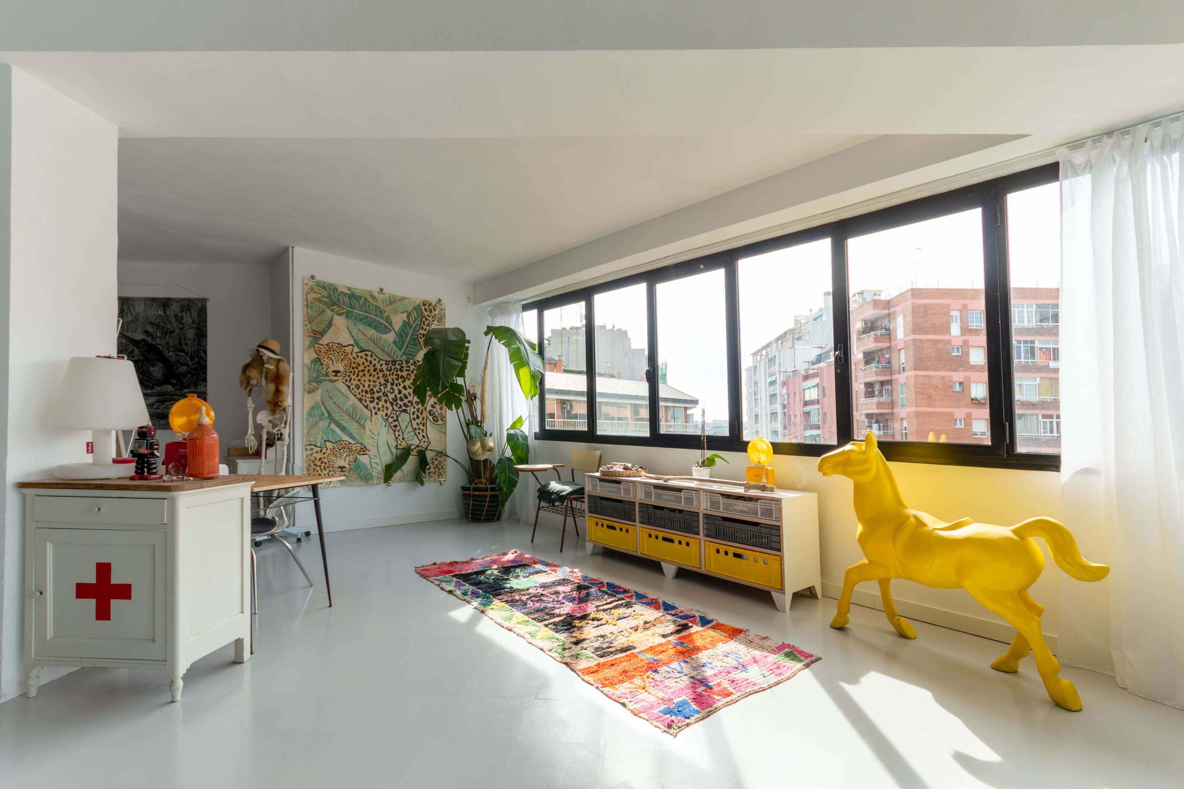 A brightly lit living space featuring large windows, a colorful rug, a yellow horse sculpture, and a mix of modern and vintage furnishings.