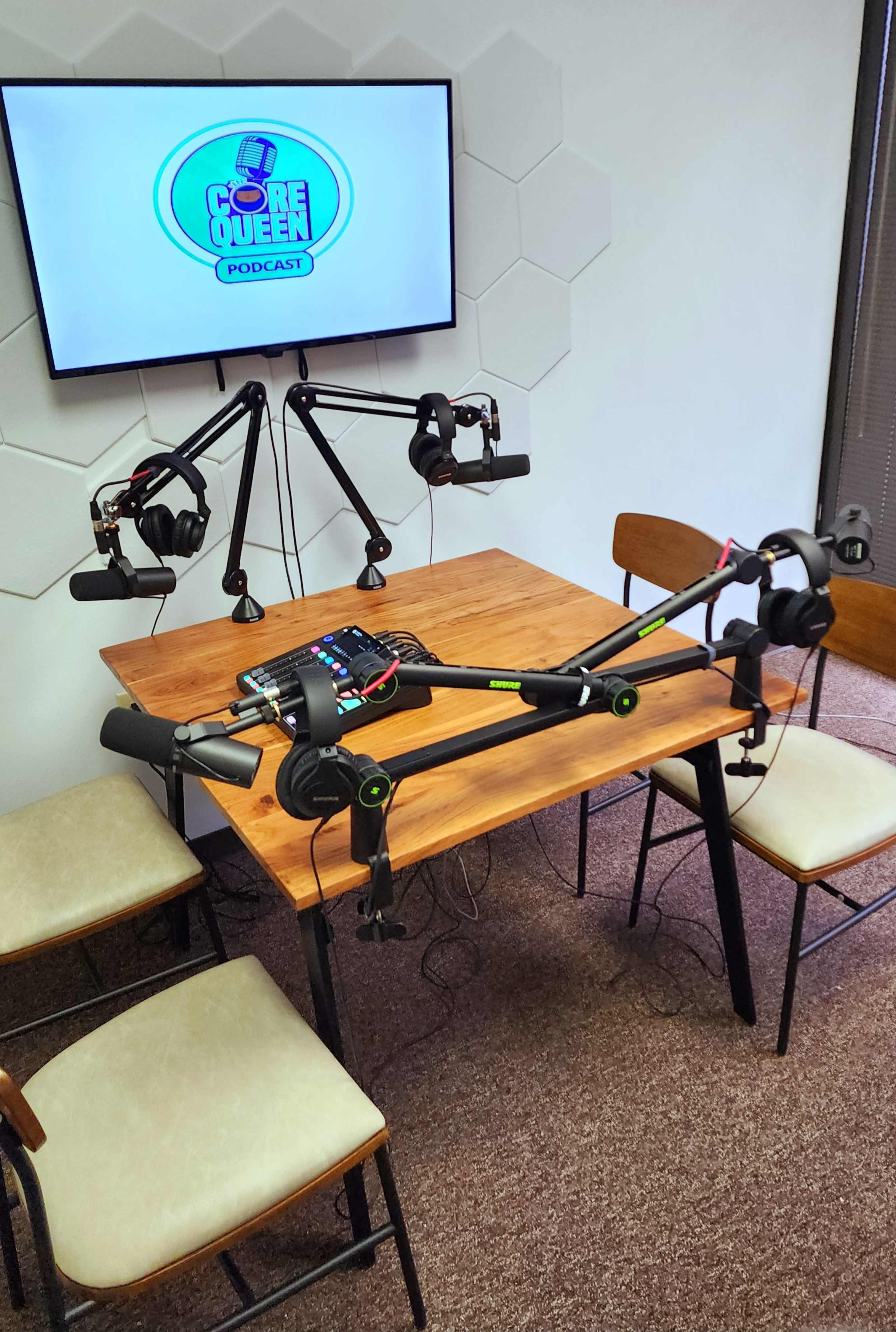 A podcast recording setup features a wooden table with four chairs, microphones on adjustable arms, and a monitor displaying "Core Queen Podcast."