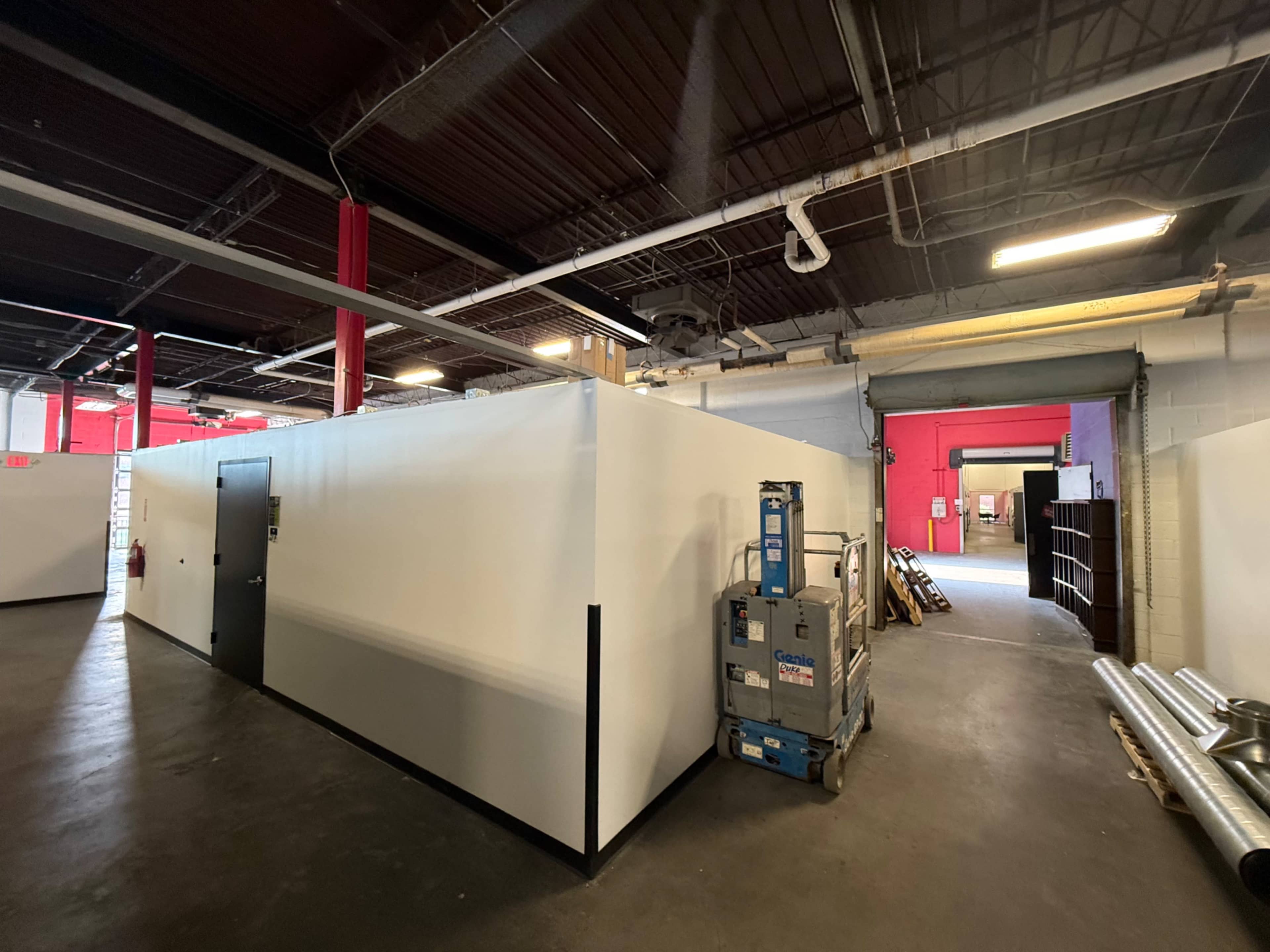 The image shows a spacious industrial area featuring white partition walls, a few pieces of construction equipment, and red accents on the walls.