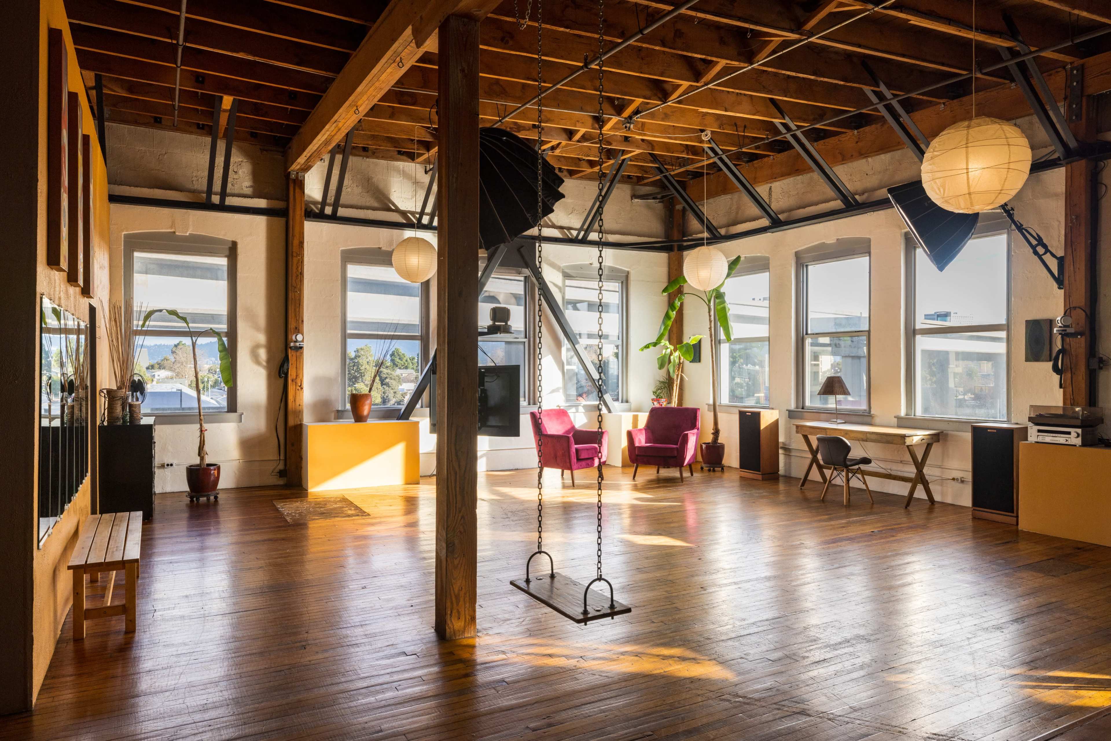 A spacious, well-lit room features wooden beams, large windows, a swing, and cozy seating areas.