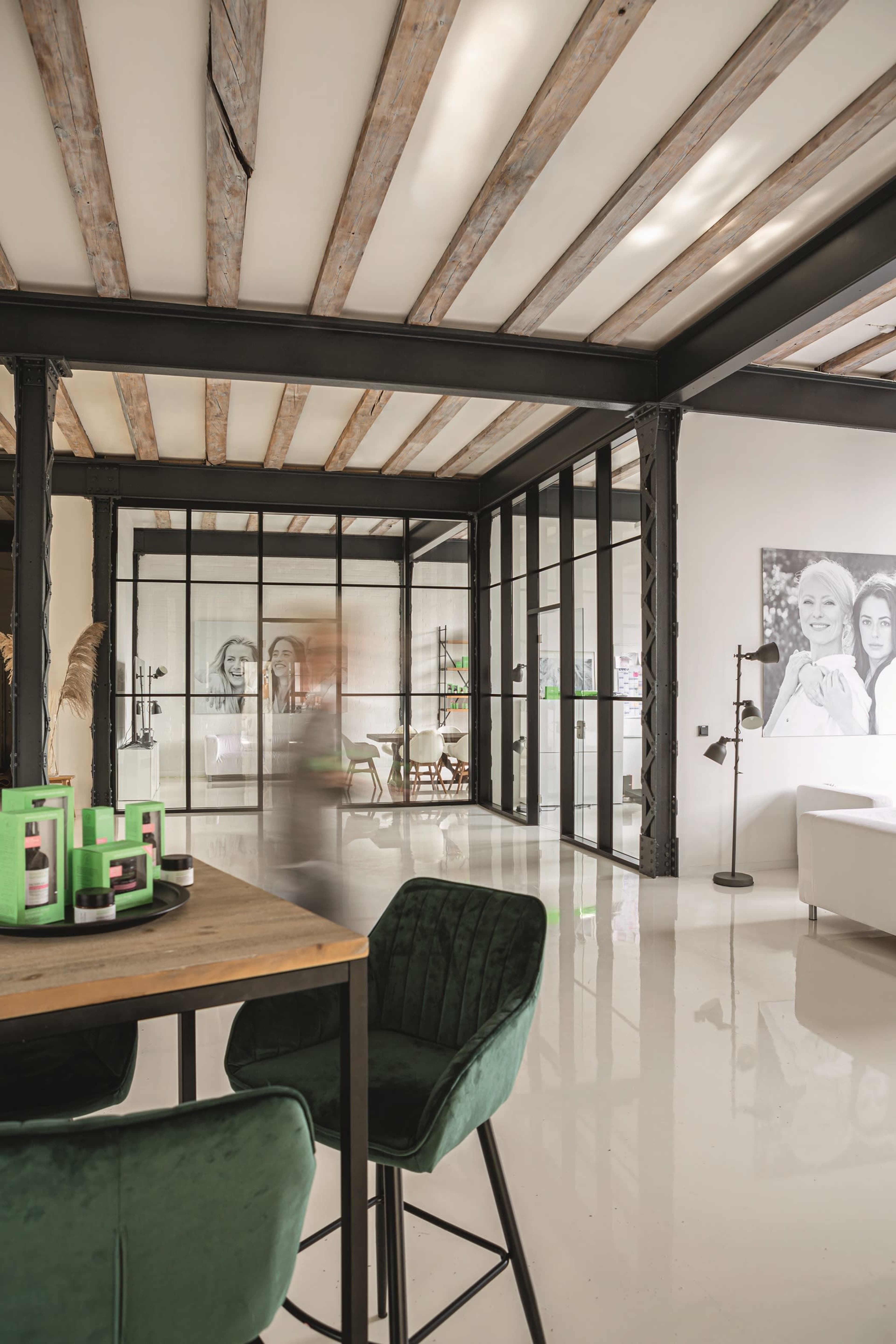 A modern open-concept space features a dining area with green velvet chairs, wooden beams on the ceiling, and large glass partitions separating different areas.