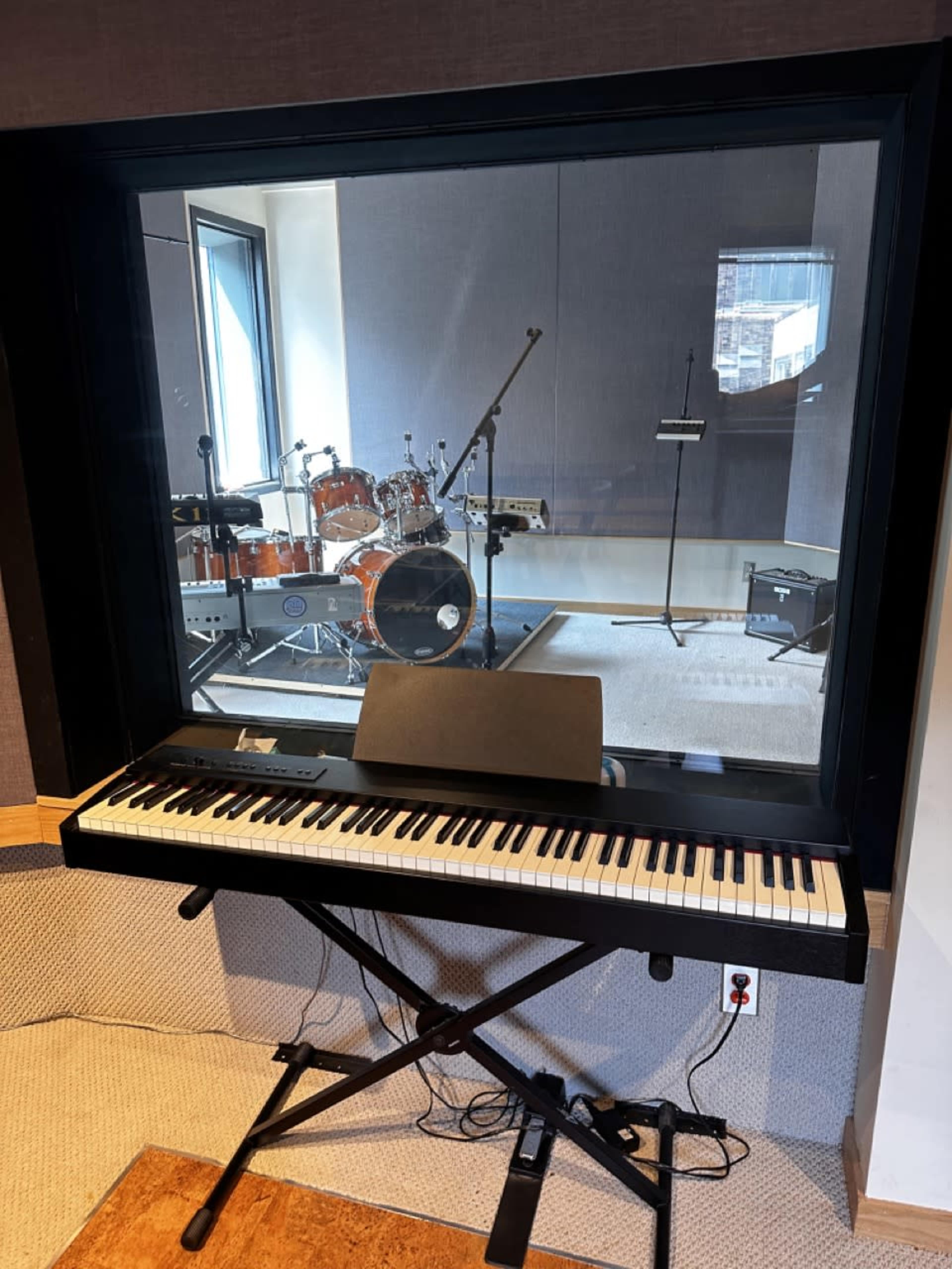 A digital piano sits in front of a large window, overlooking a recording studio with a drum set and microphone inside.