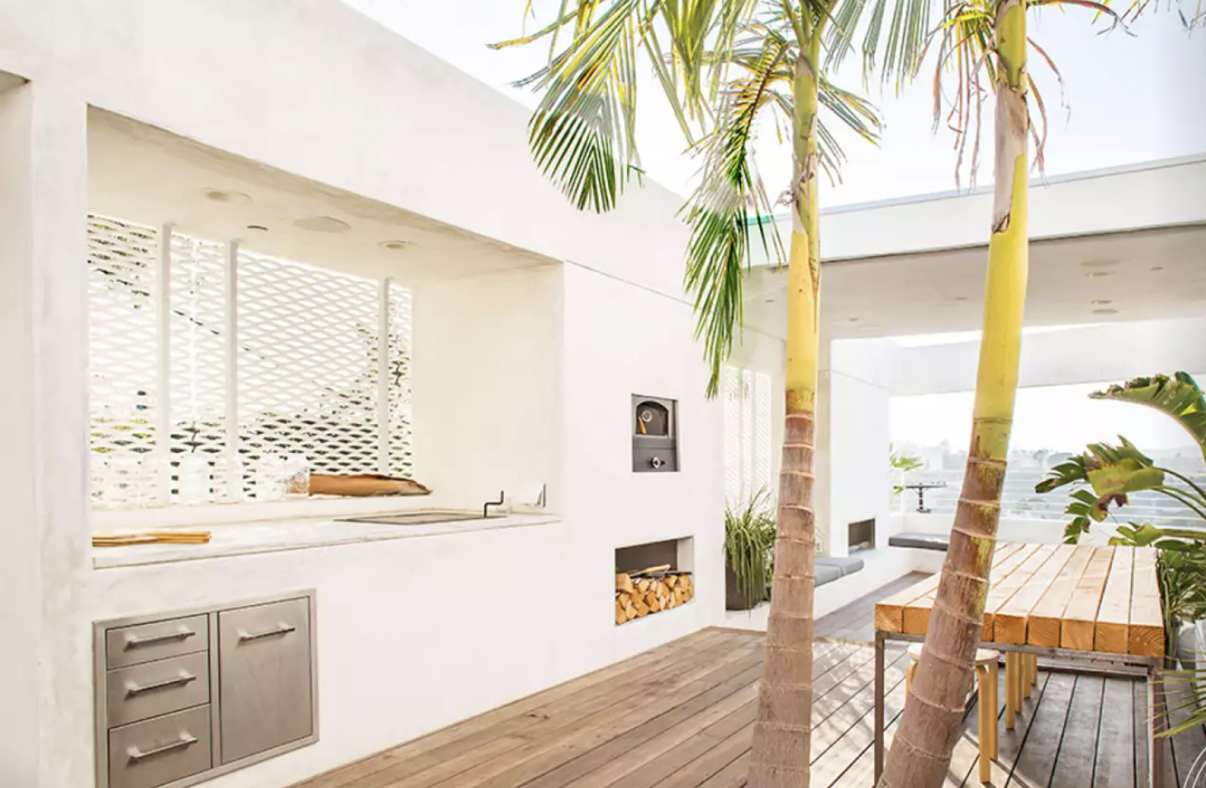 A modern outdoor space featuring a minimalist kitchen area with a grill, a wooden dining table, and tall palm trees.