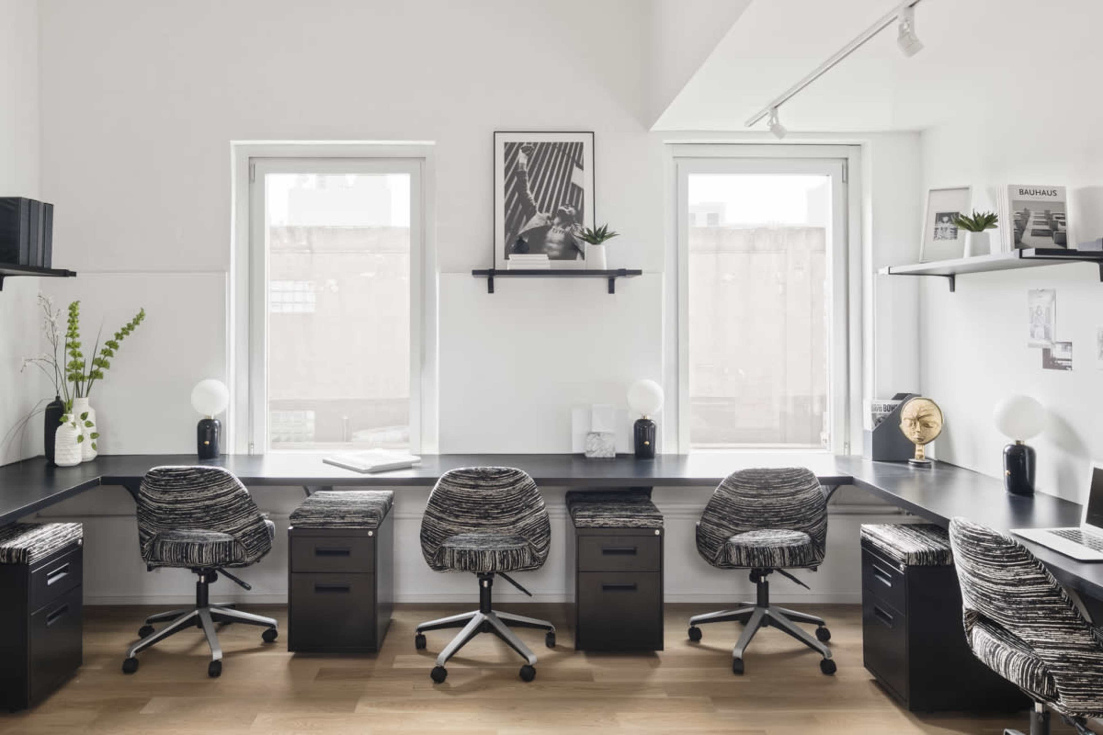 A modern office space features four patterned swivel chairs along a sleek black desk, with large windows and minimalistic decor.