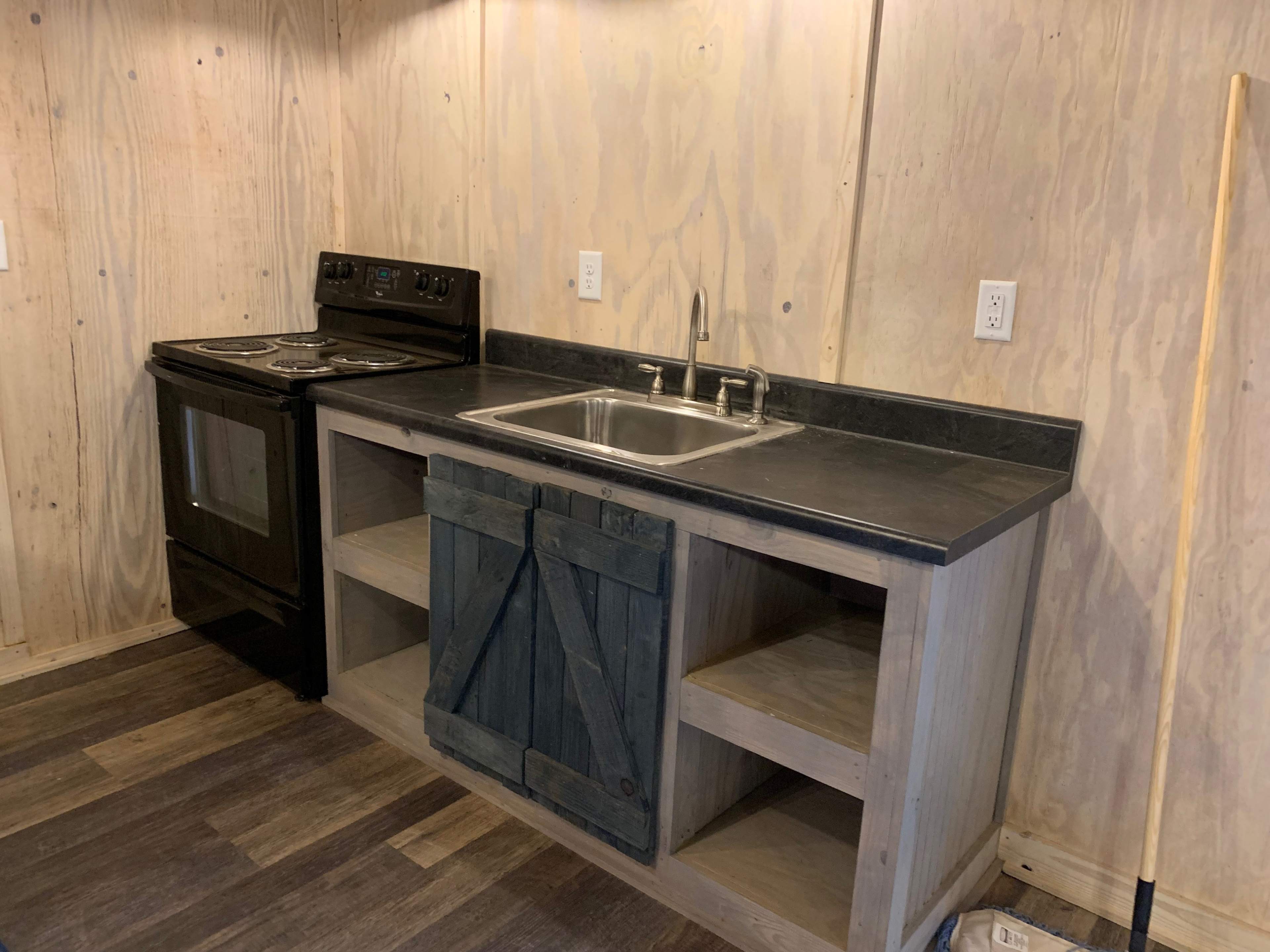 The image shows a small kitchen area featuring a black stove and oven next to a stainless steel sink, set on a wooden countertop with open shelving beneath.