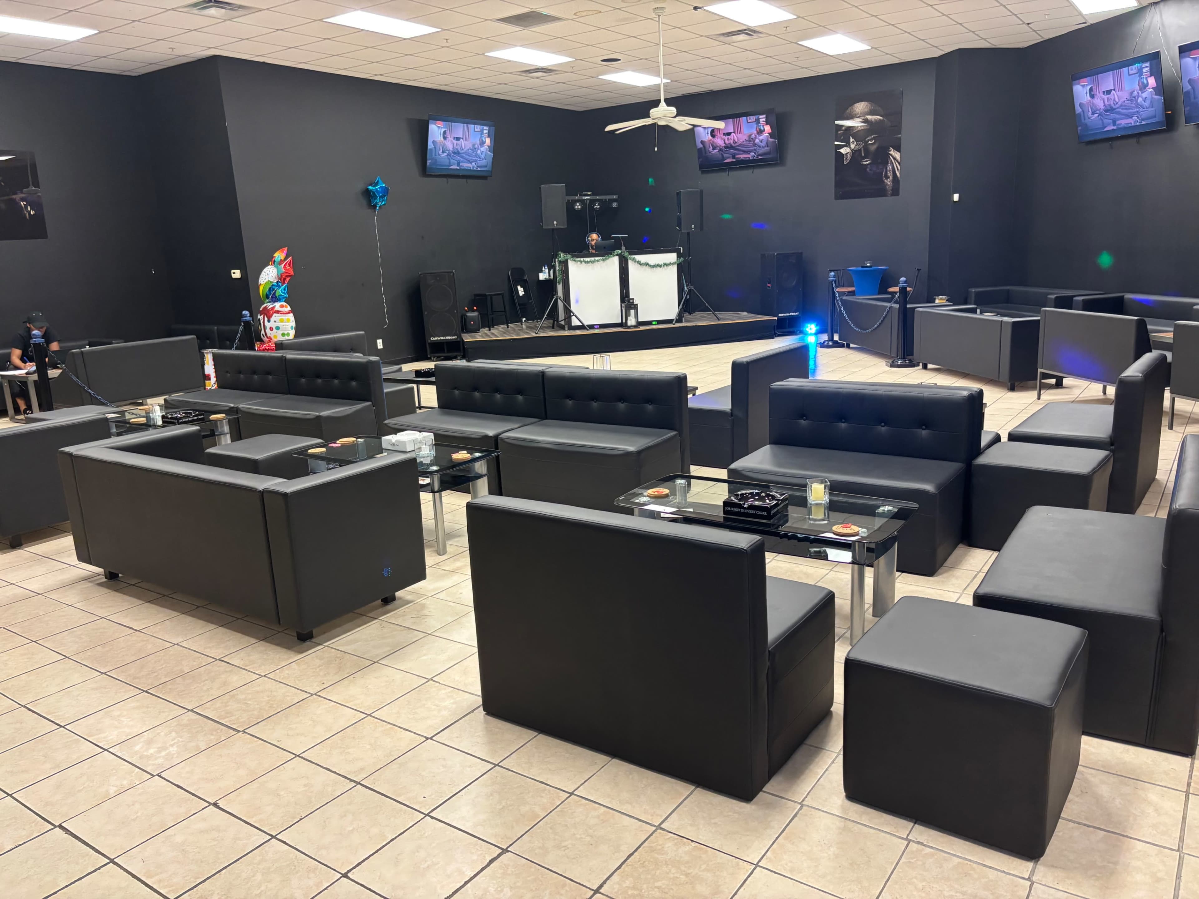 The image shows a spacious lounge area with black furniture, a stage at the front, and two televisions mounted on the walls.