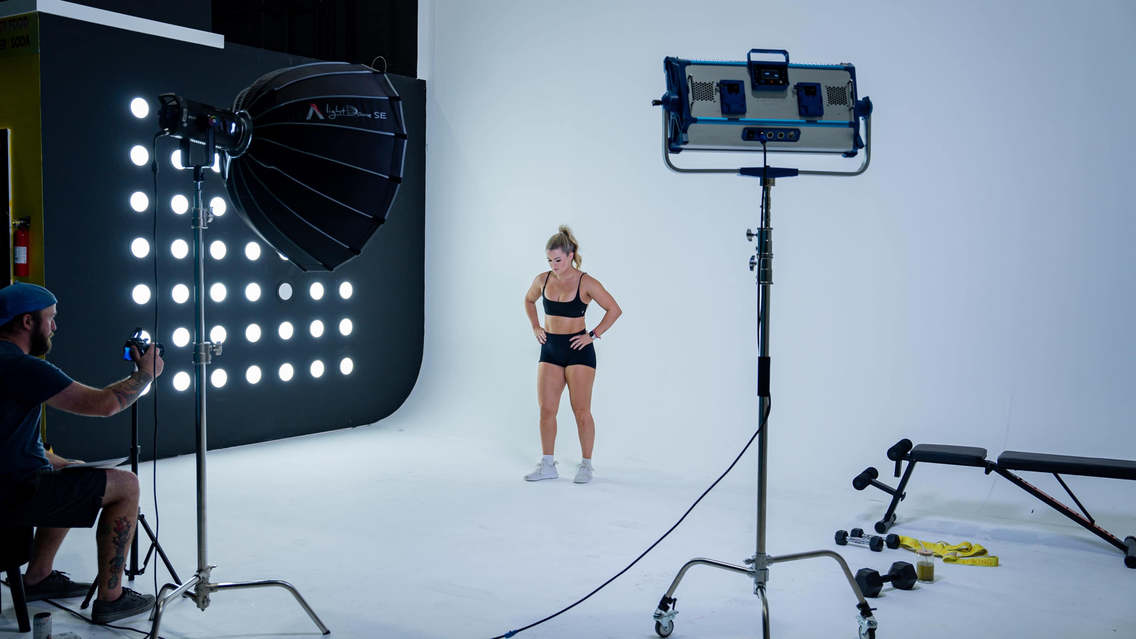A fitness model poses in a studio setting with professional lighting and camera equipment.