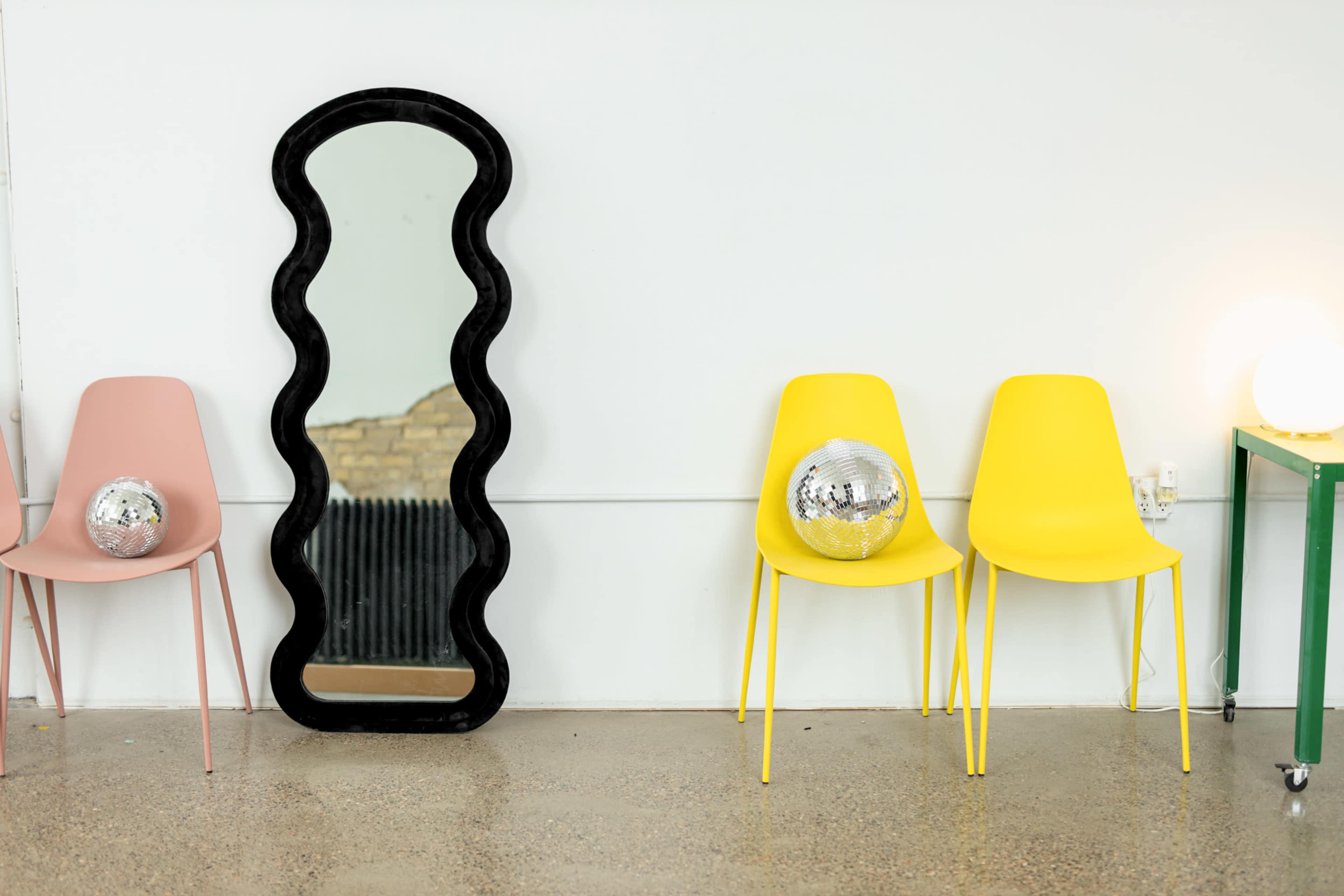 A black wavy mirror stands against a white wall, flanked by a pink chair with a disco ball and a yellow chair with another disco ball.