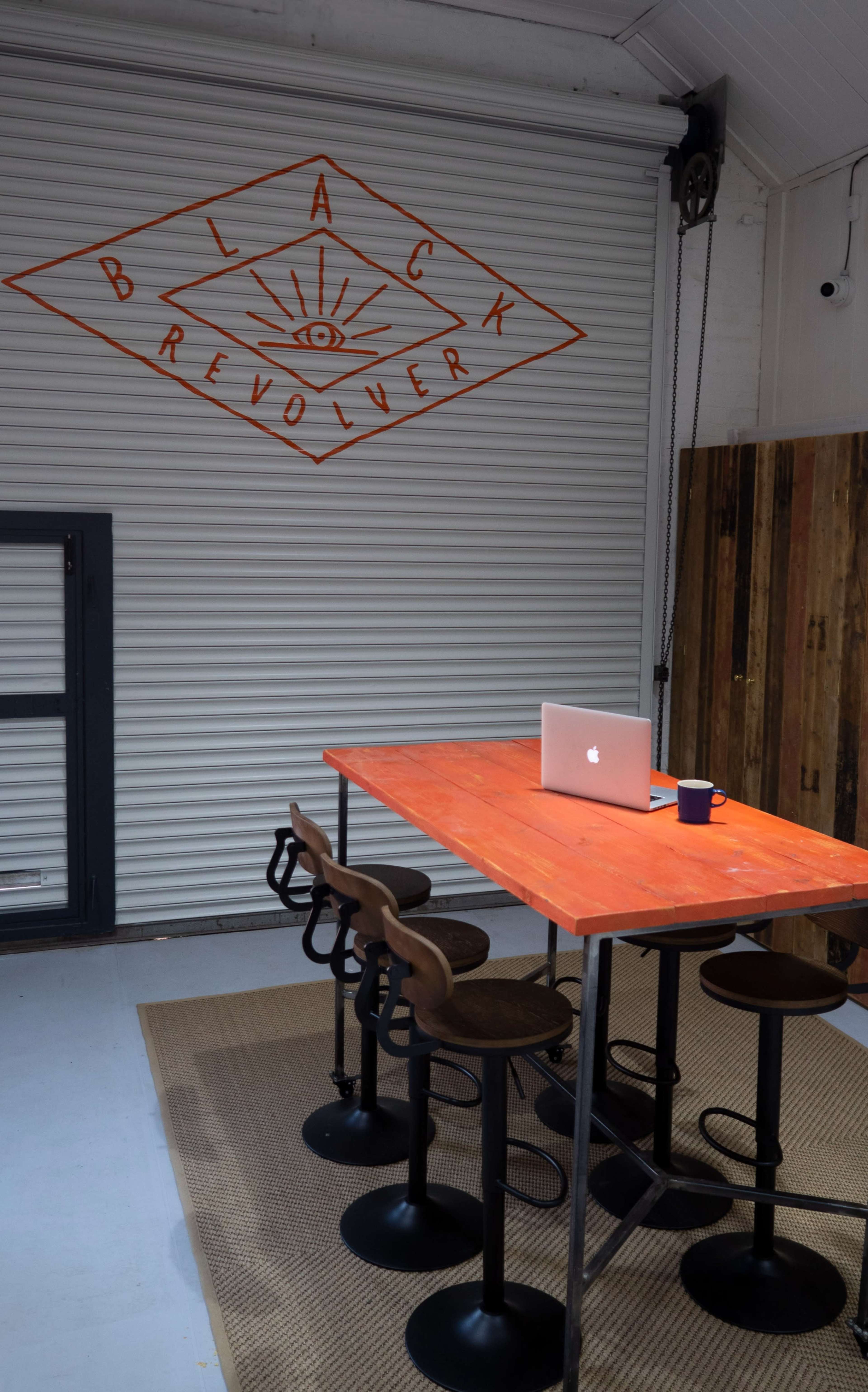 A simple workspace features a vibrant orange table with four black stools, a laptop, and a coffee mug, against a backdrop of a closed garage door with an orange graphic design.